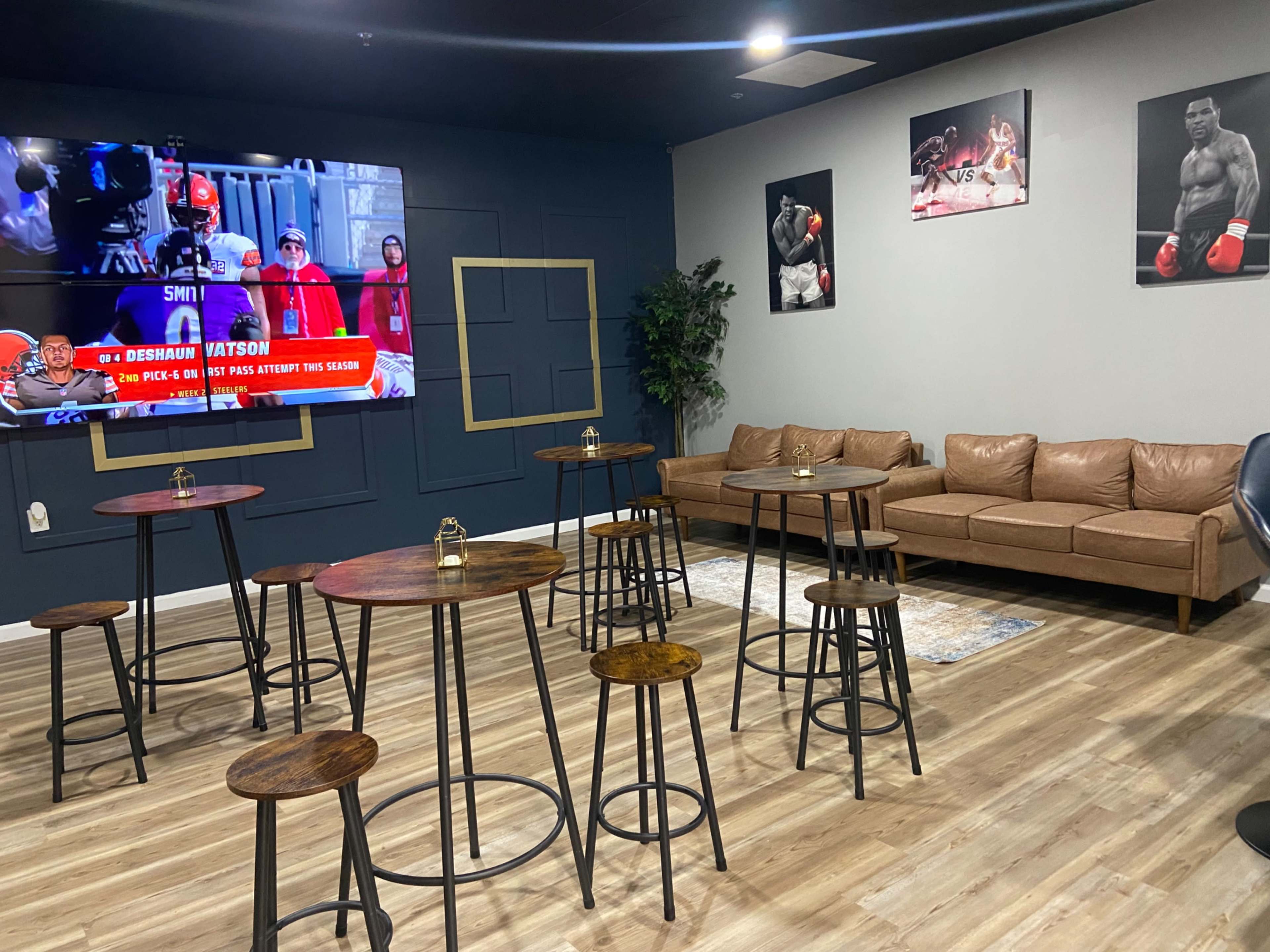 The image shows a spacious lounge area featuring several high tables and stools, a leather sofa, and wall-mounted sports-themed artwork, along with a large television displaying a sports event.