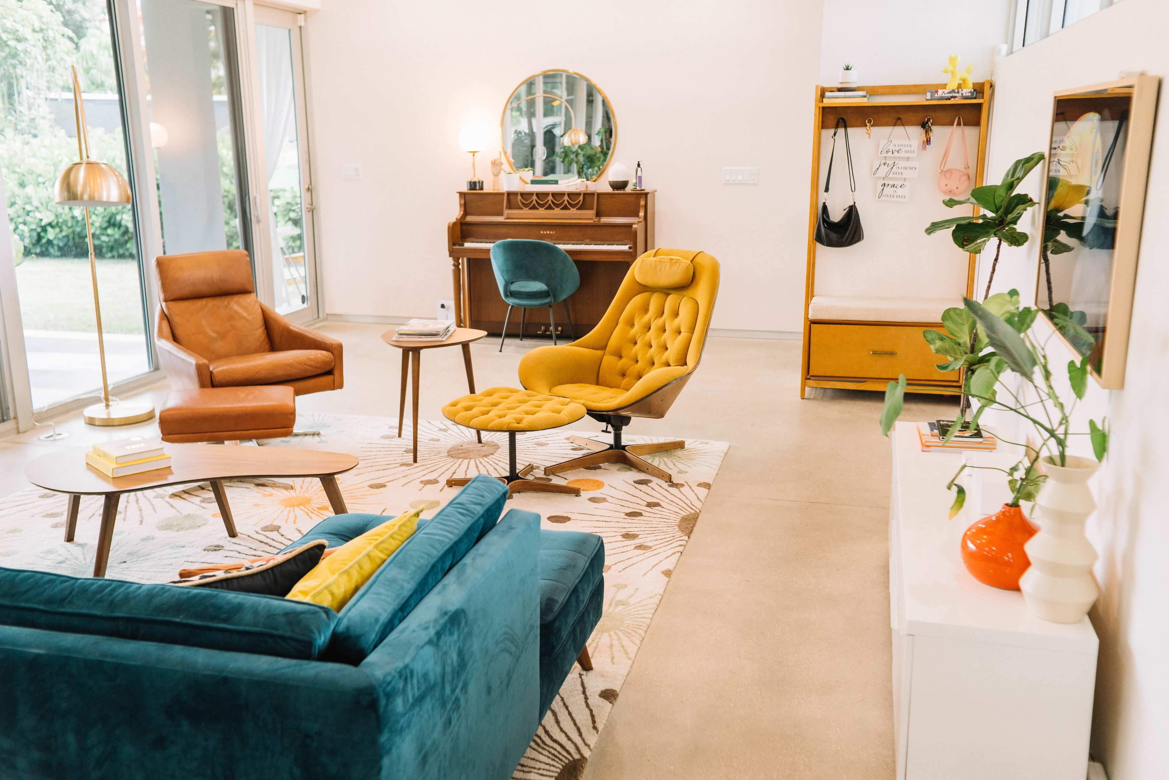 A modern living room features a blue couch, an orange chair, a yellow lounge chair, a round coffee table, and a piano, all arranged around a patterned rug.