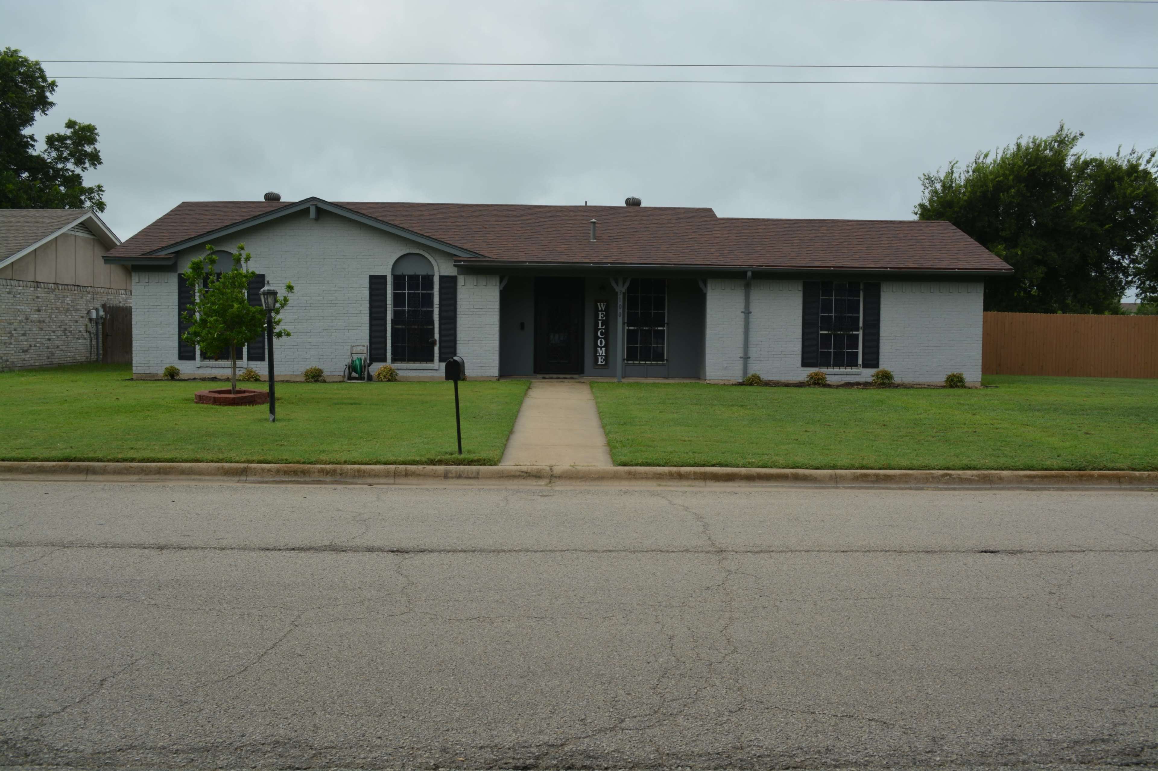 A single-story, light gray house with a dark brown roof, situated on a quiet street, features a manicured lawn and a paved walkway leading to the front door.