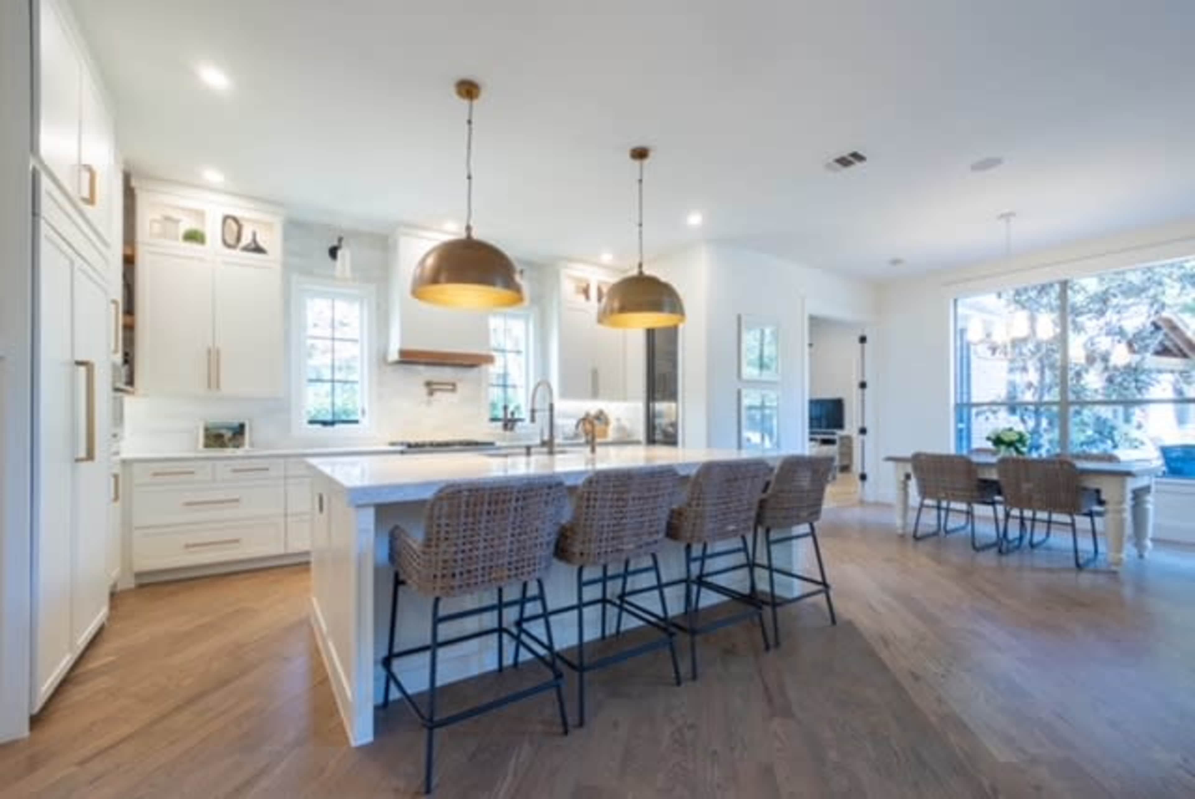 A modern kitchen with white cabinetry, a large island with bar stools, and pendant lighting, adjacent to a dining area with large windows.