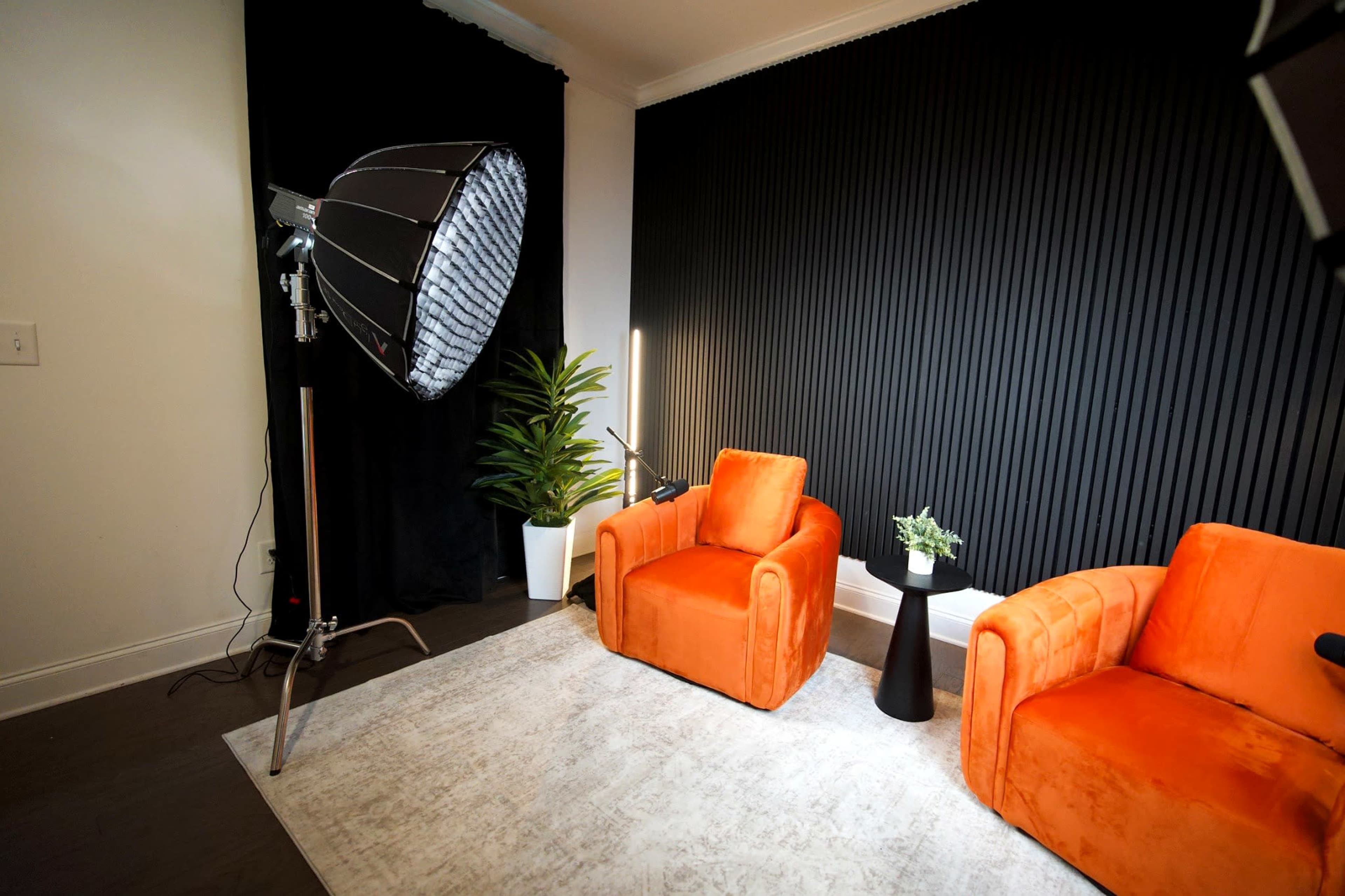 The image features a well-lit interior scene with two orange armchairs, a small black table, and a potted plant against a black, ribbed wall.