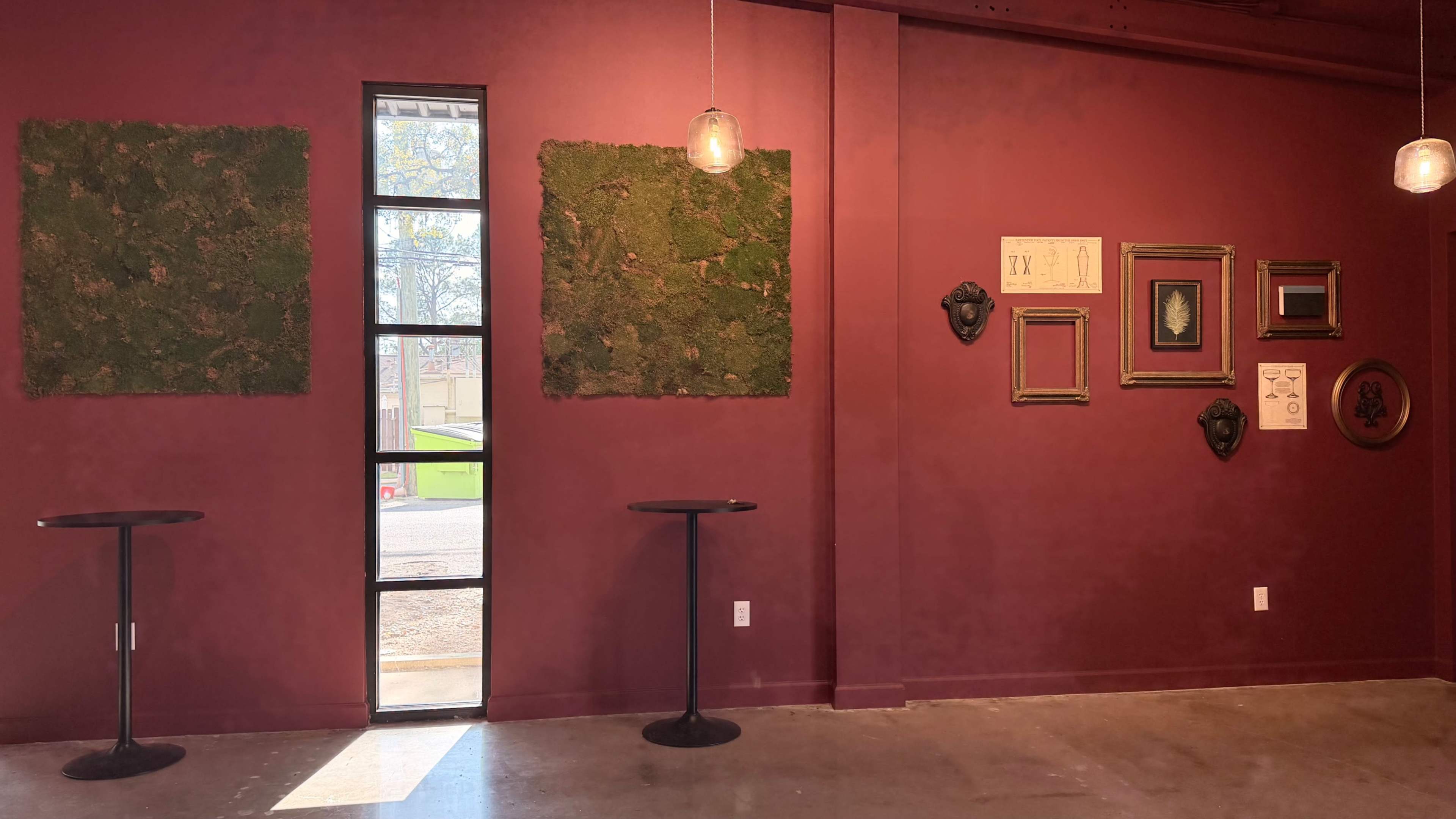 The image shows a wall with two green moss panels, a tall window opening onto the street, and various framed art pieces arranged decoratively on a deep red wall.