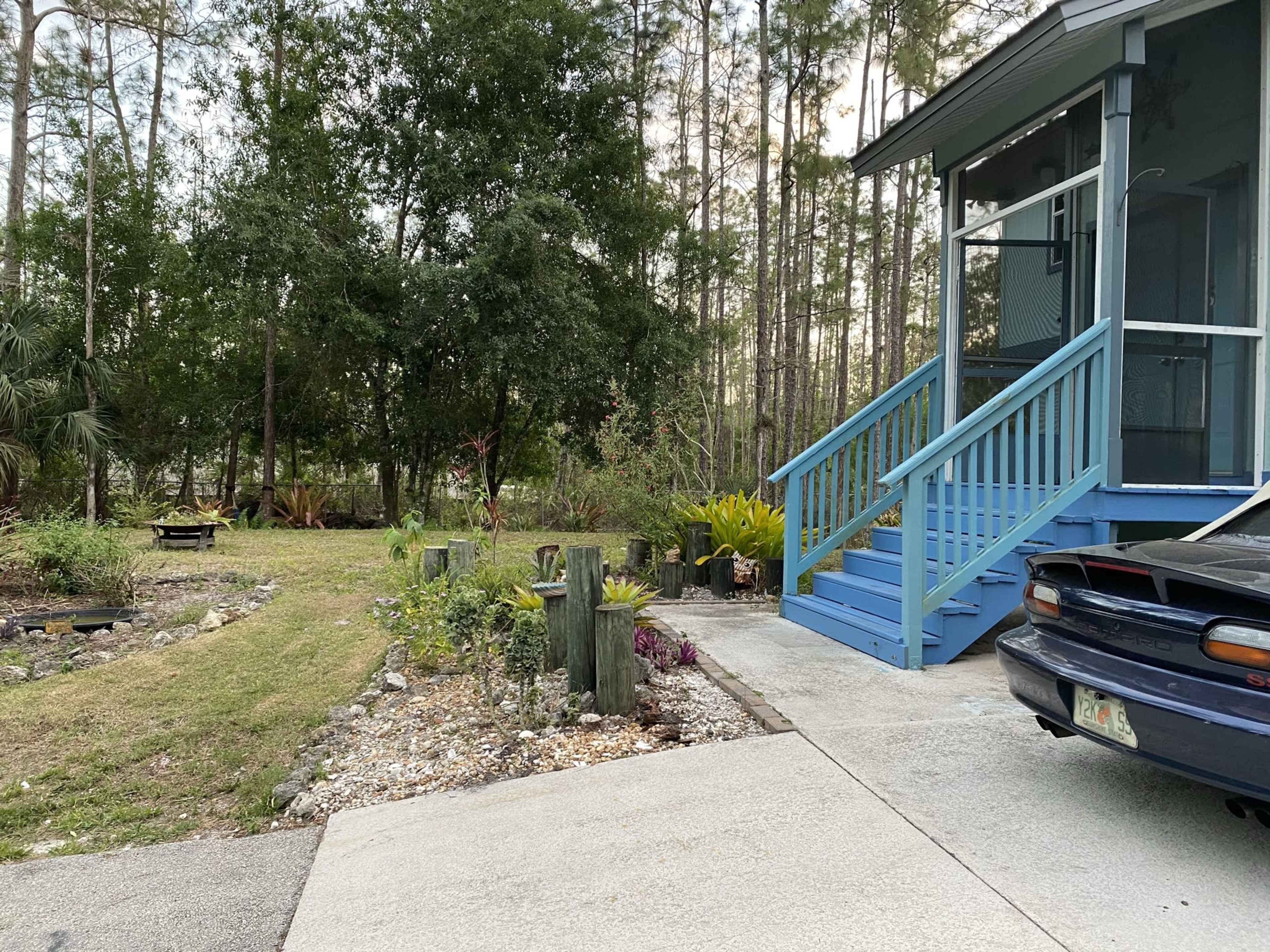 A blue house with a porch and steps is situated next to a gravel driveway that leads to a landscaped yard with trees in the background.