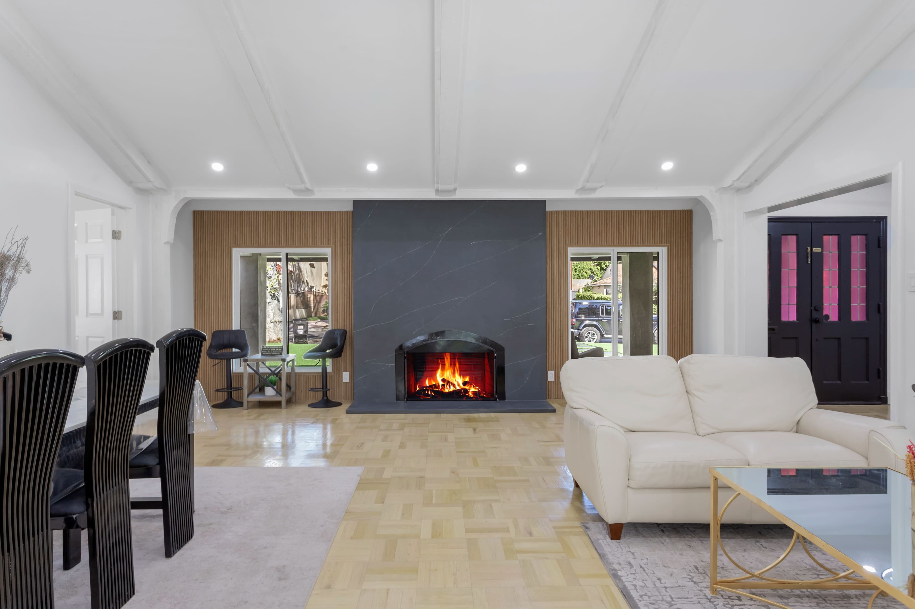 A modern living room with a fireplace, a cream-colored sofa, a dining area with black chairs, and large windows illuminating the space.