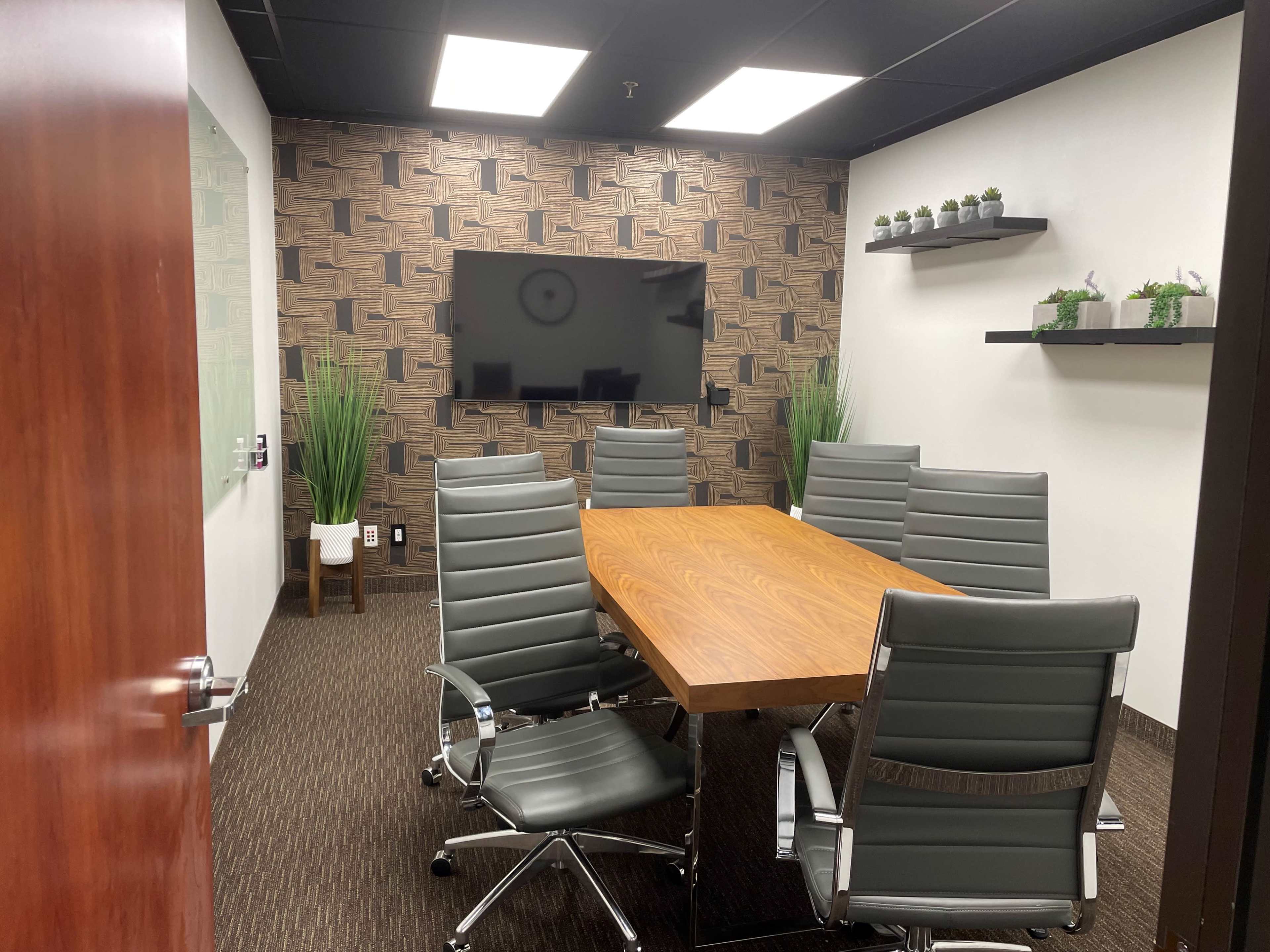 A conference room features a wooden table surrounded by six gray leather chairs, with a wall-mounted television and decorative plants on shelves.