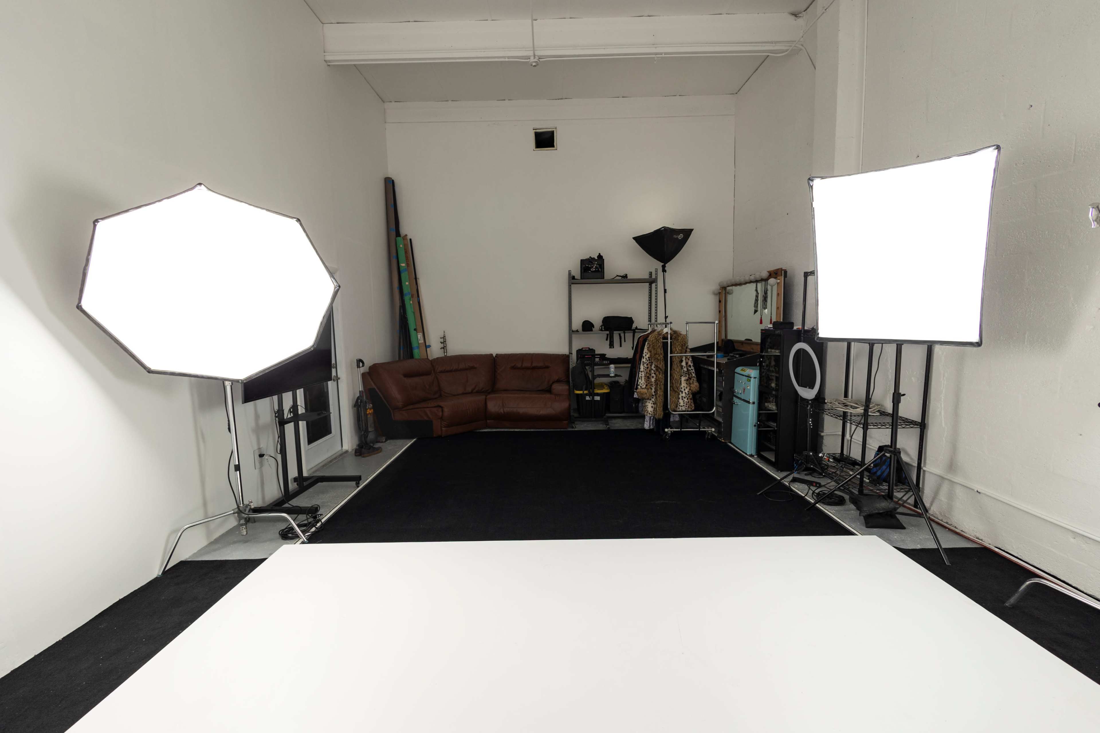 A photography studio with a white backdrop, two large softbox lights, a brown couch, and various props arranged on shelves.