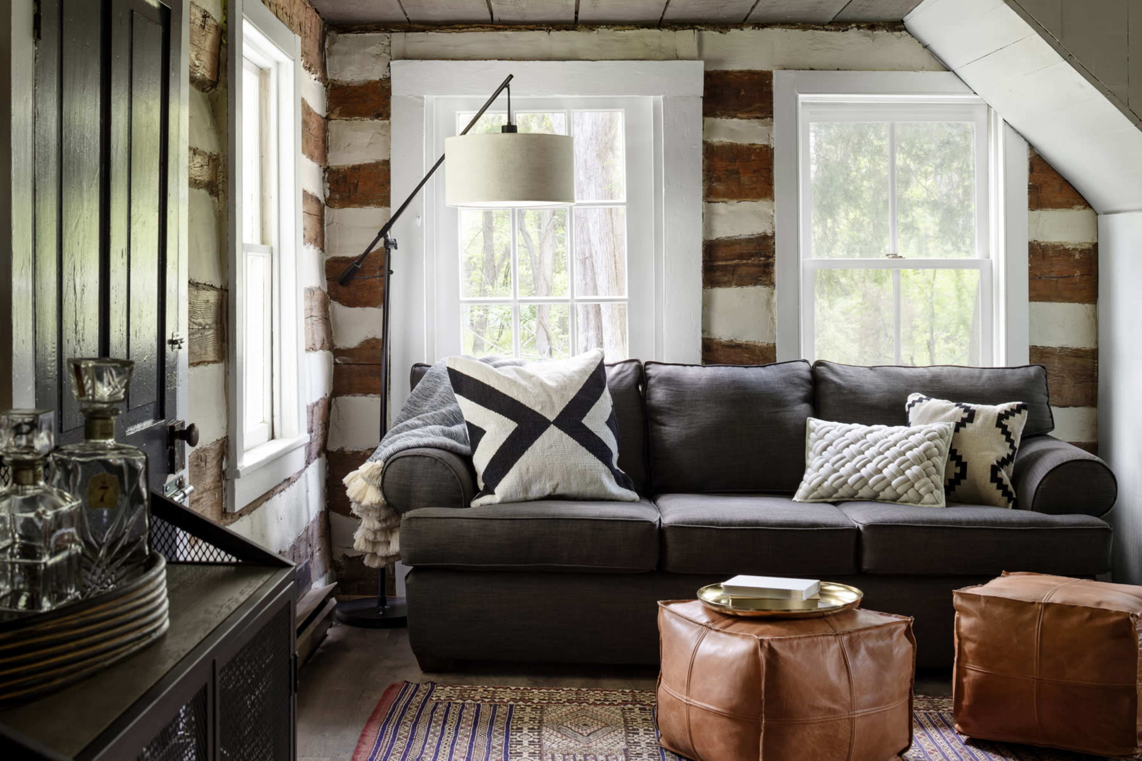 A cozy living room featuring a gray sofa adorned with decorative pillows, flanked by windows and rustic wood-paneled walls, alongside a round leather ottoman.