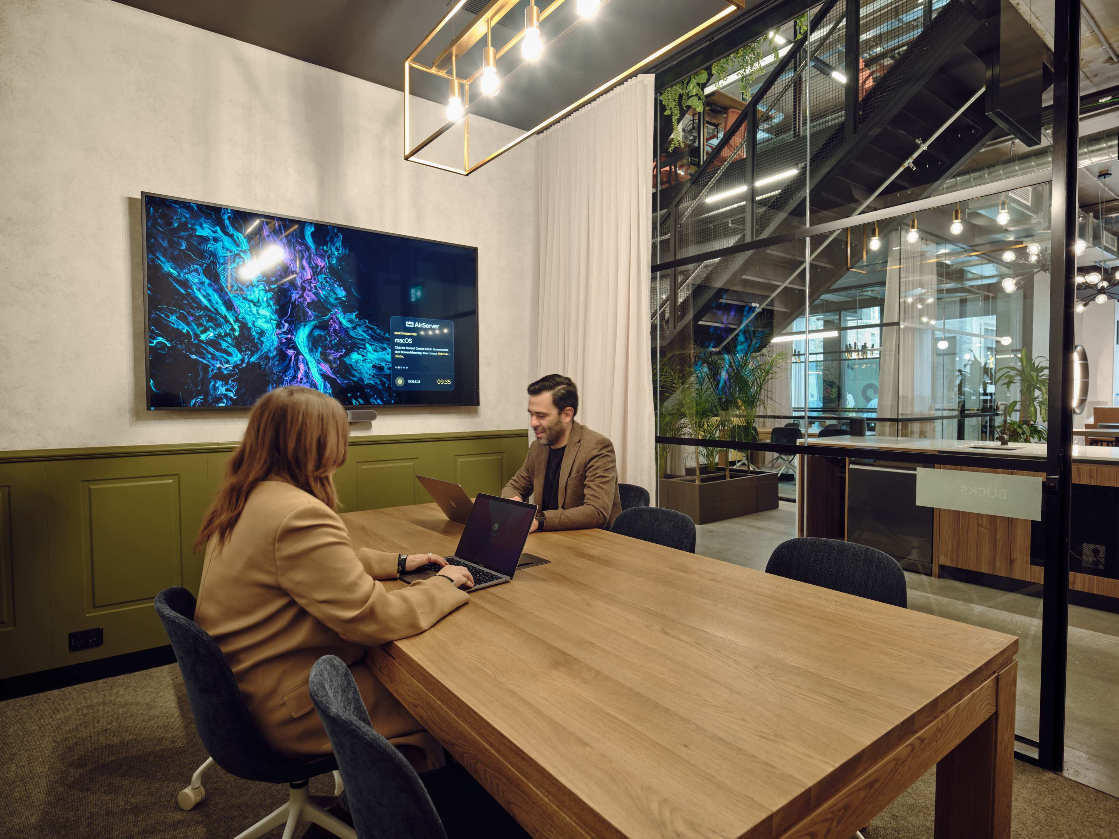 Two people are seated at a wooden table in a modern conference room with a large screen displaying colorful graphics.