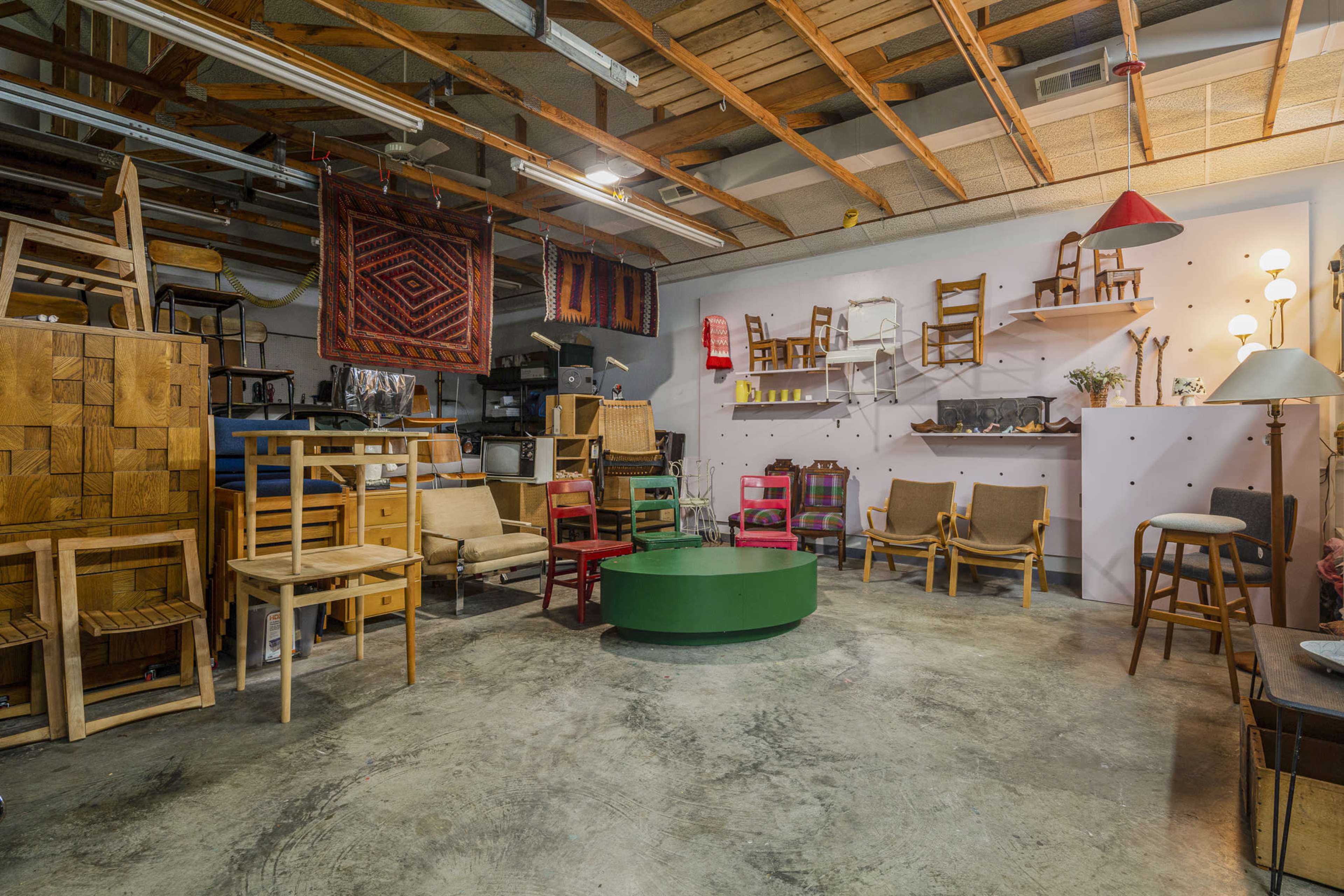 A cluttered workshop or studio space containing various wooden furniture pieces, including chairs and tables, with artwork hanging on the walls.
