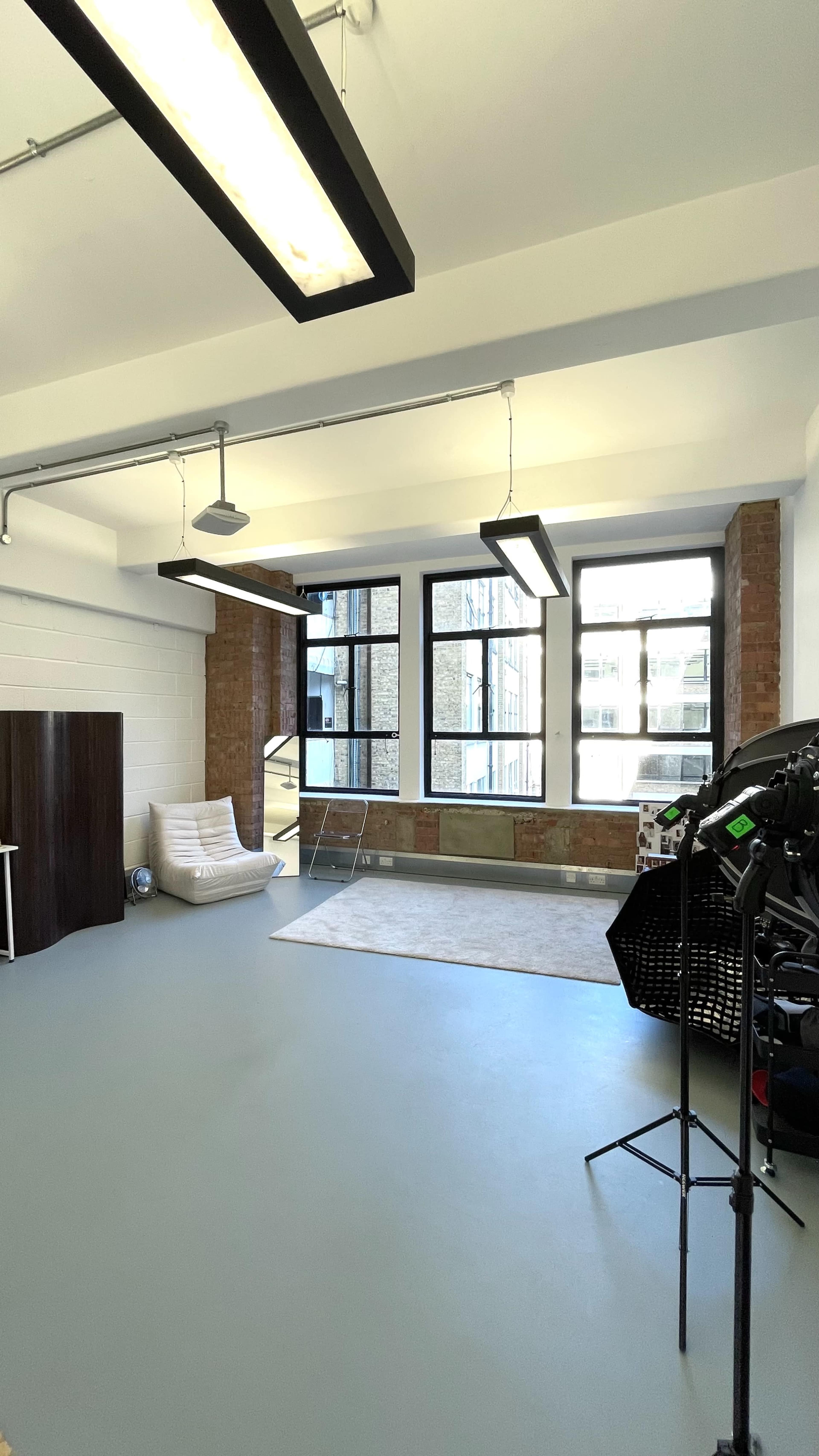 The image shows a spacious, well-lit room featuring large windows, concrete walls, and a minimalist design with furniture including a couch and a light stand.