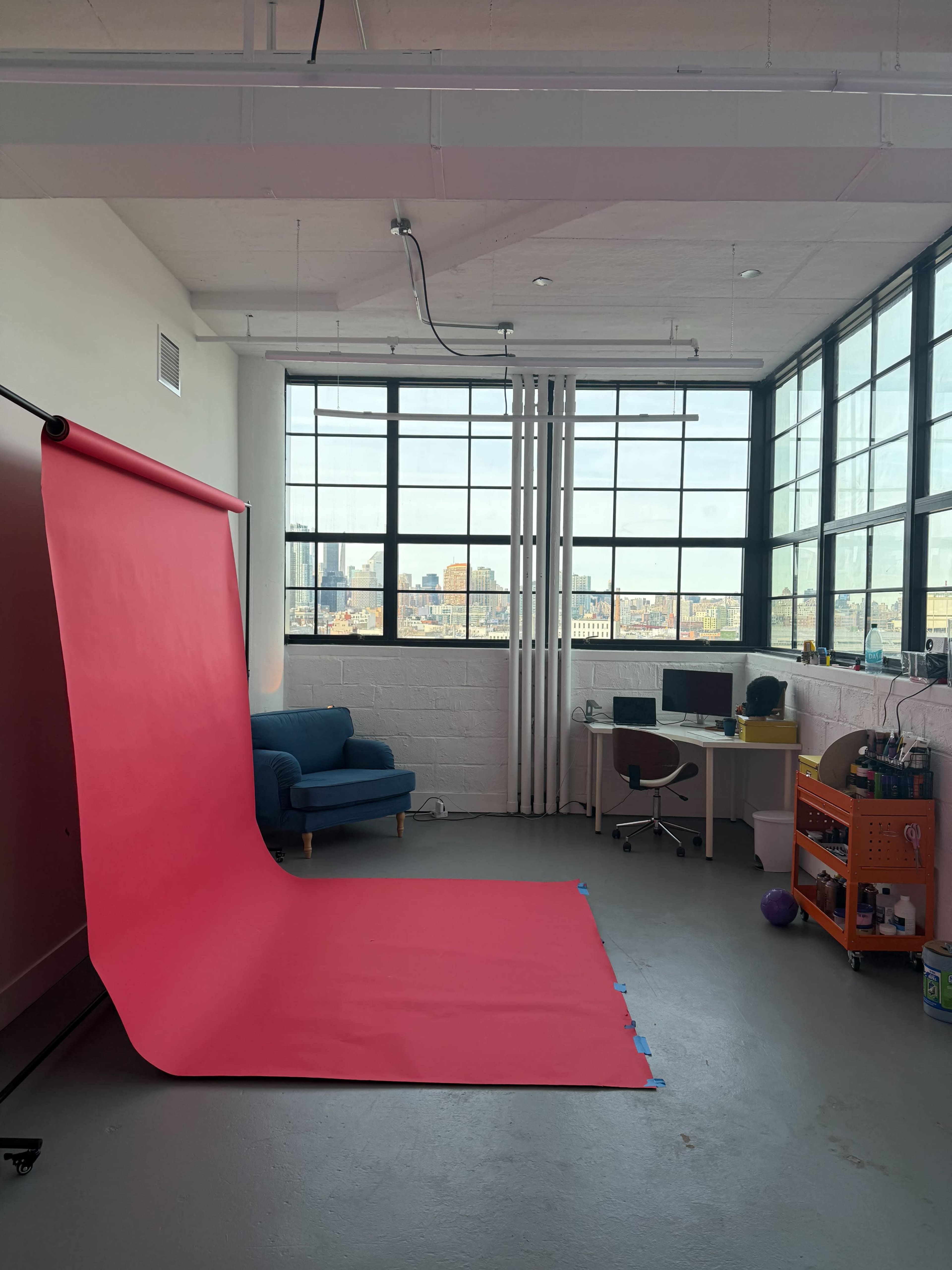 The image shows a bright studio space featuring a large pink backdrop, a blue couch, a desk with a computer, and a rolling cart against a wall of large windows.