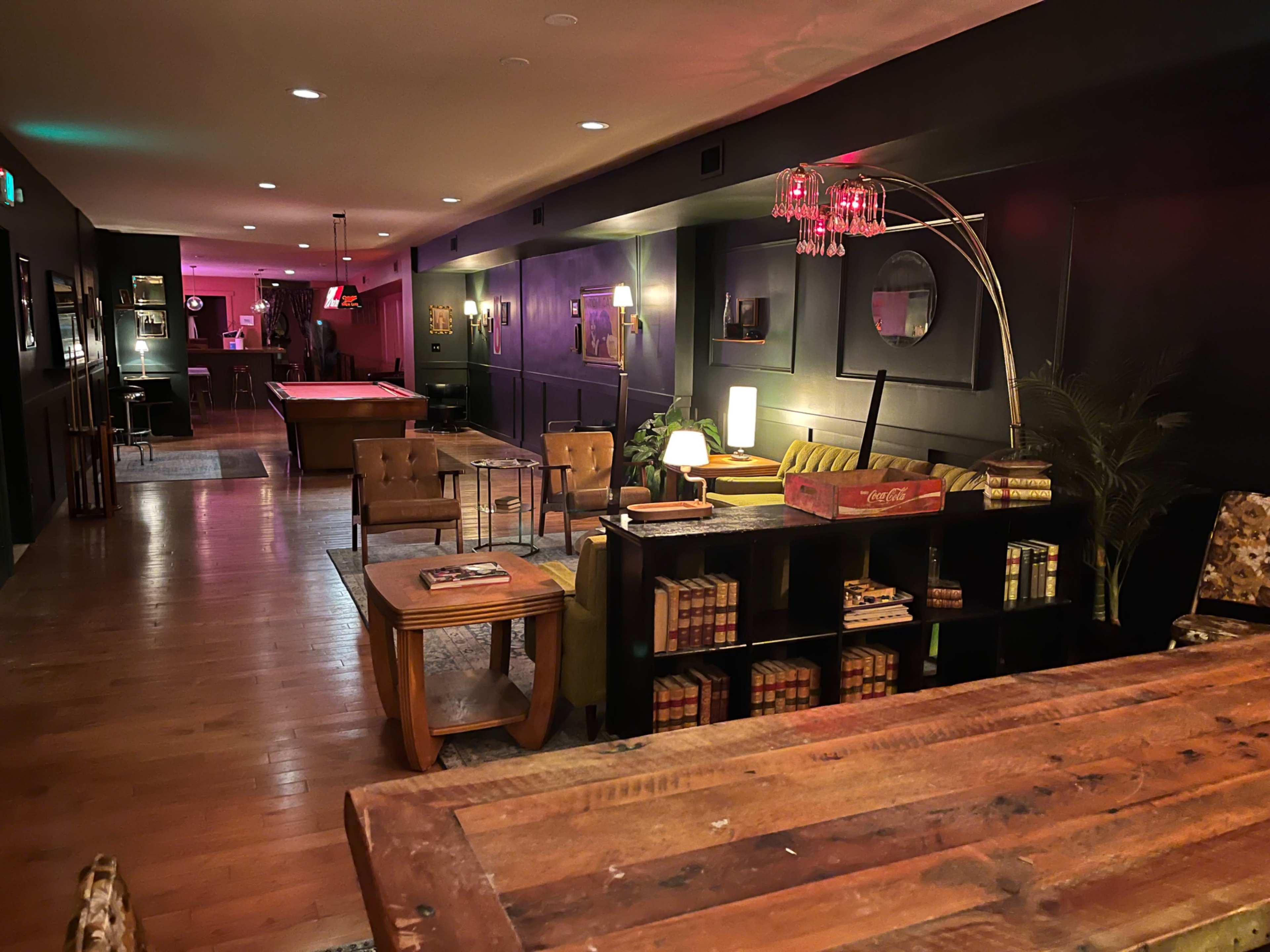 The scene shows a dimly lit lounge area featuring a mix of seating arrangements, a pool table in the background, and decorative elements like books and a lamp on a shelf.