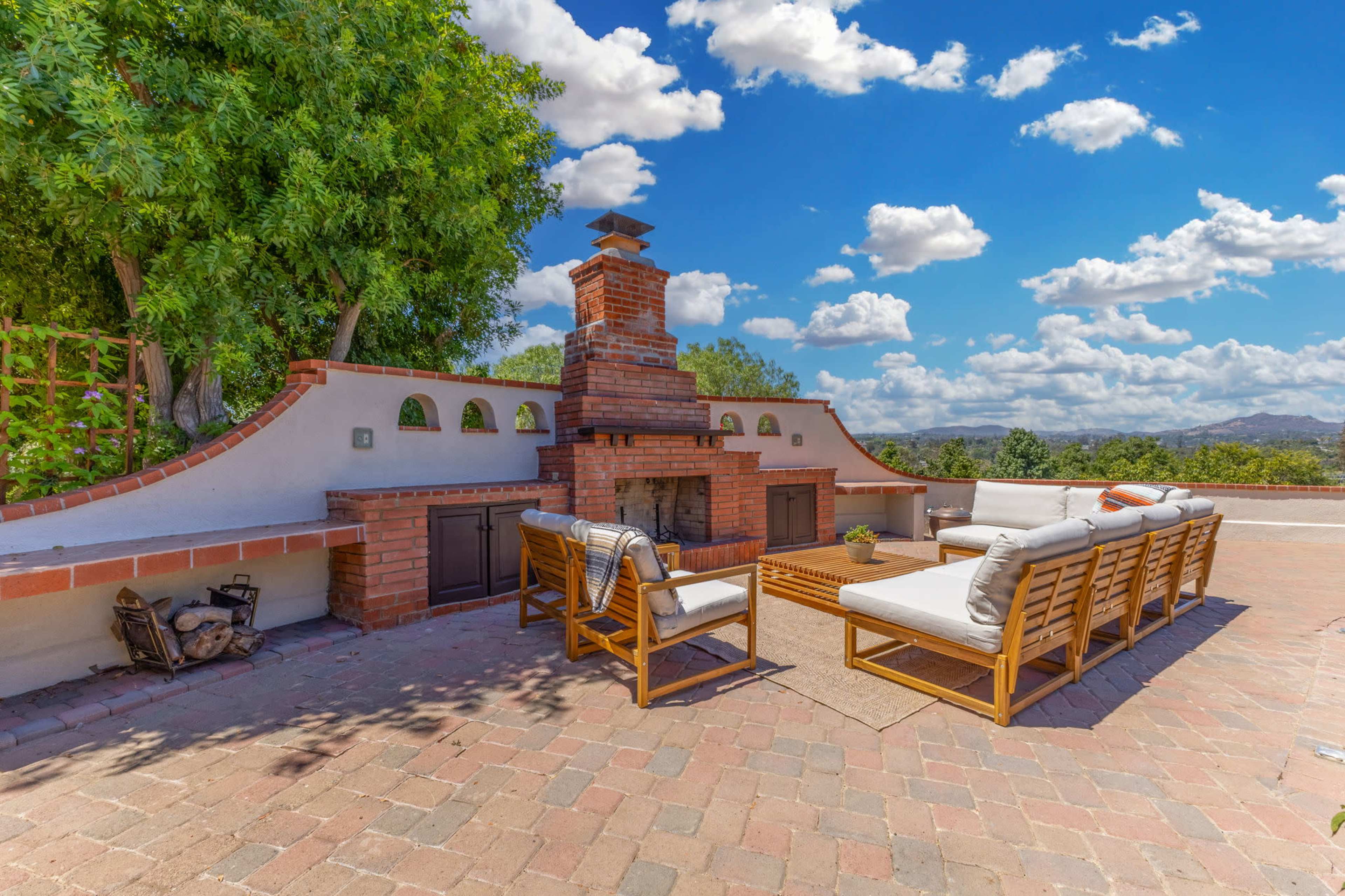 The image shows an outdoor patio area with a brick fireplace, surrounded by seating arranged on a stone-paved surface under a blue sky with scattered clouds.