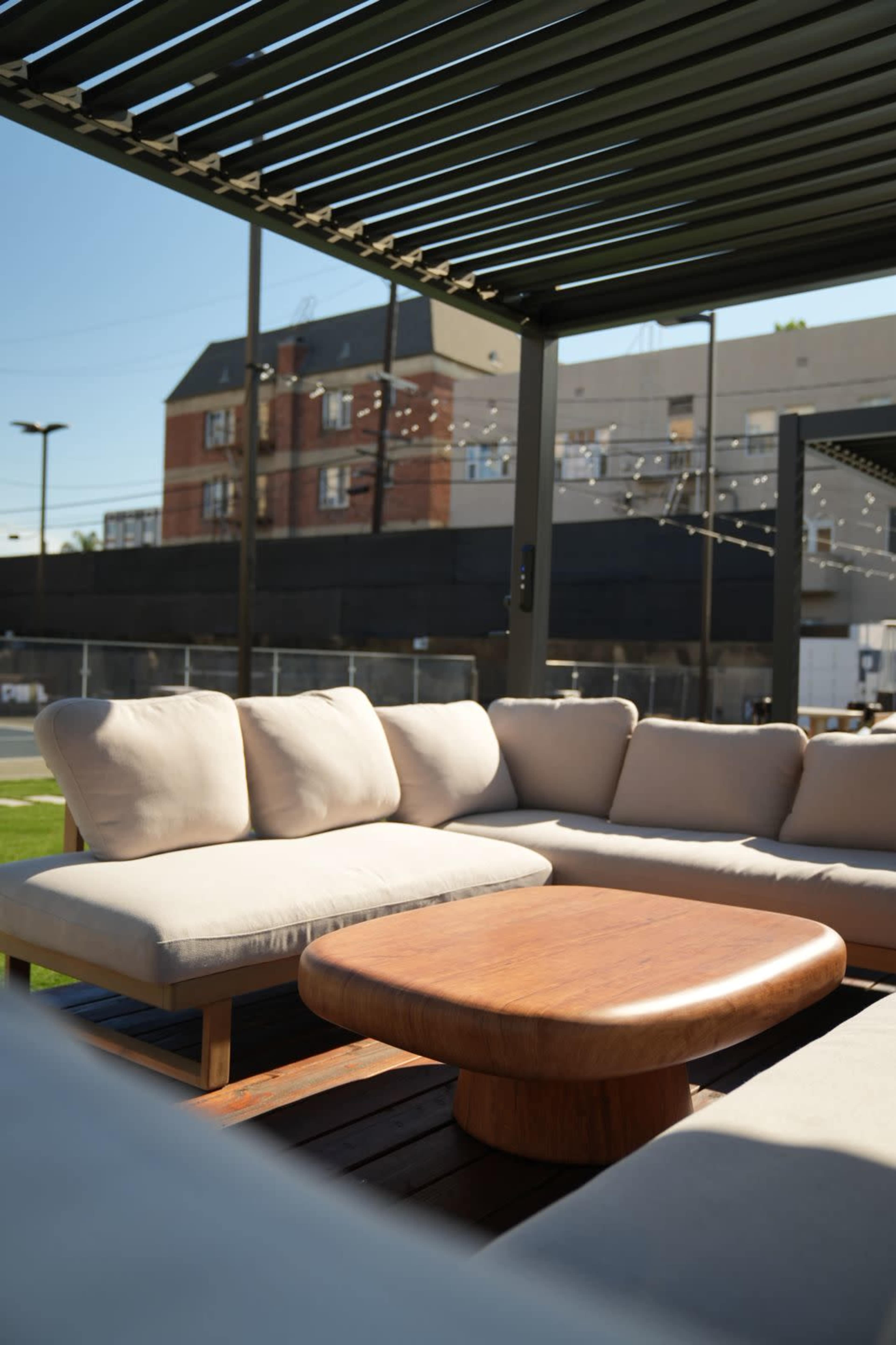 The image shows a modern outdoor seating arrangement with a beige sectional sofa and a round wooden coffee table under a shaded pergola.