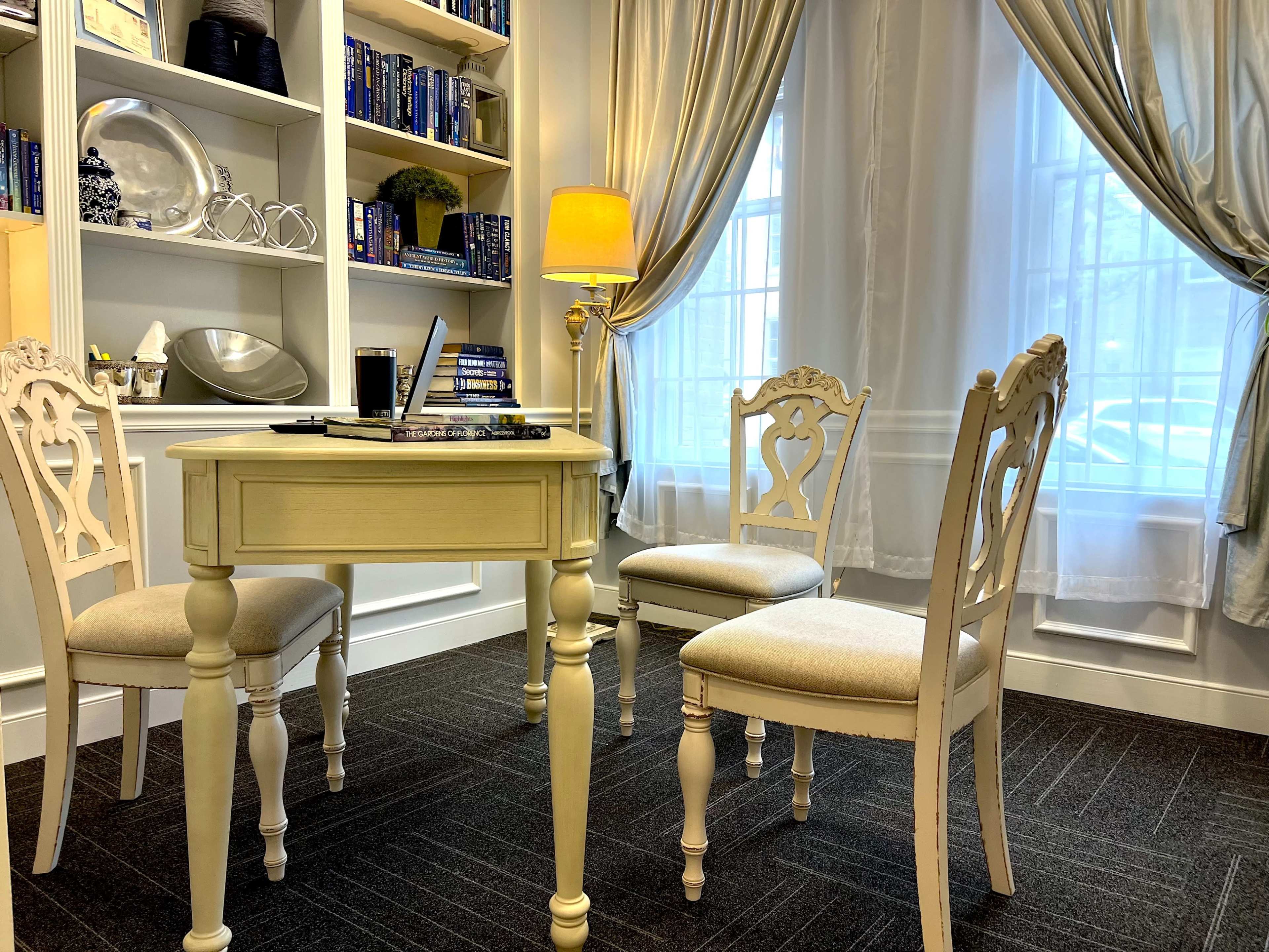 A small study area features a light-colored table surrounded by three chairs, with bookshelves in the background and a lamp illuminating the space.