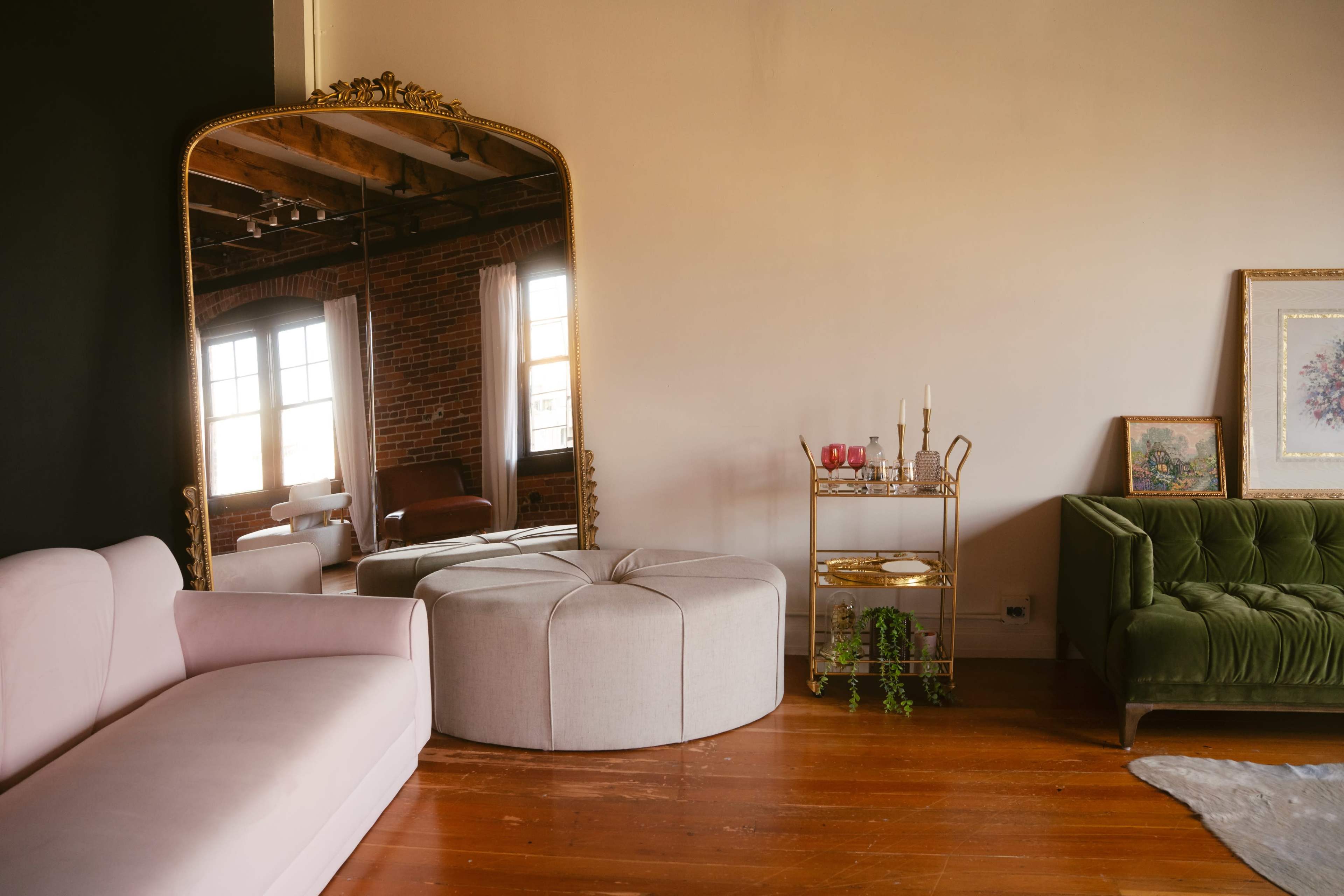 The image shows a stylish interior with a large ornate mirror, a round fabric ottoman, a pink sofa, and a green velvet couch, accompanied by a small gold bar cart with decorative items.