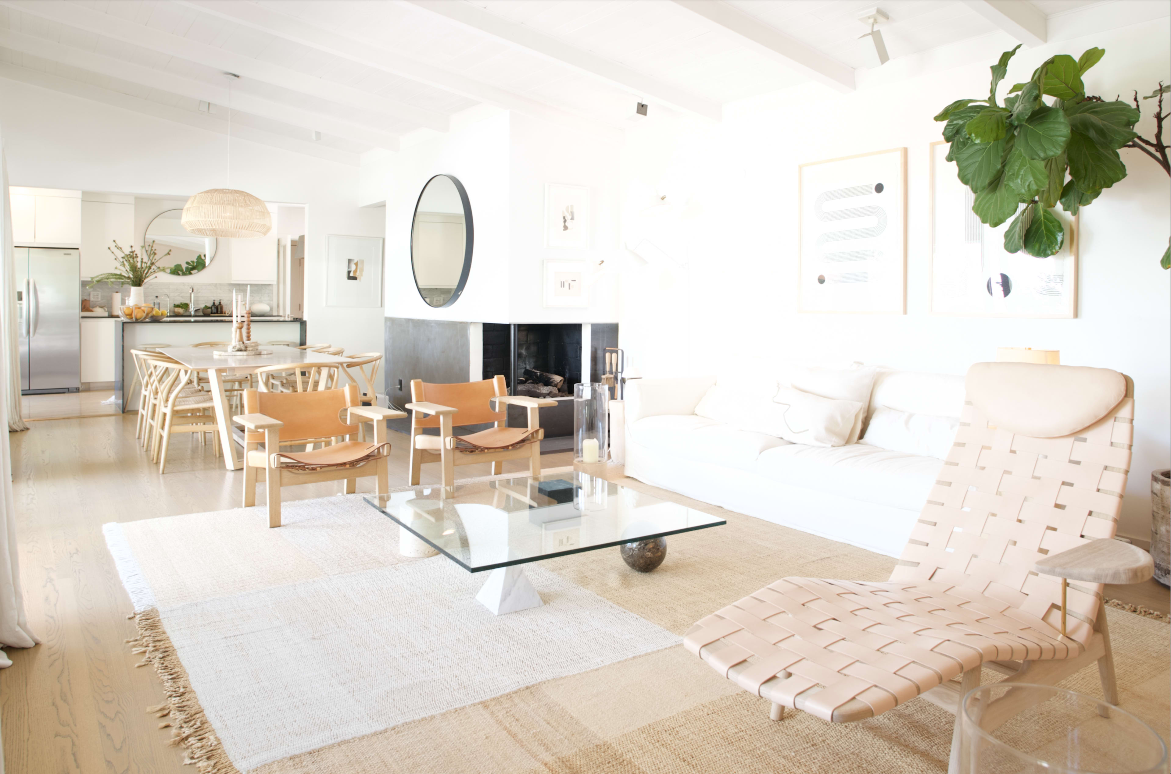 A bright, modern living room features a white sofa, wooden chairs, a glass coffee table, and a dining area with a circular mirror and a fireplace.