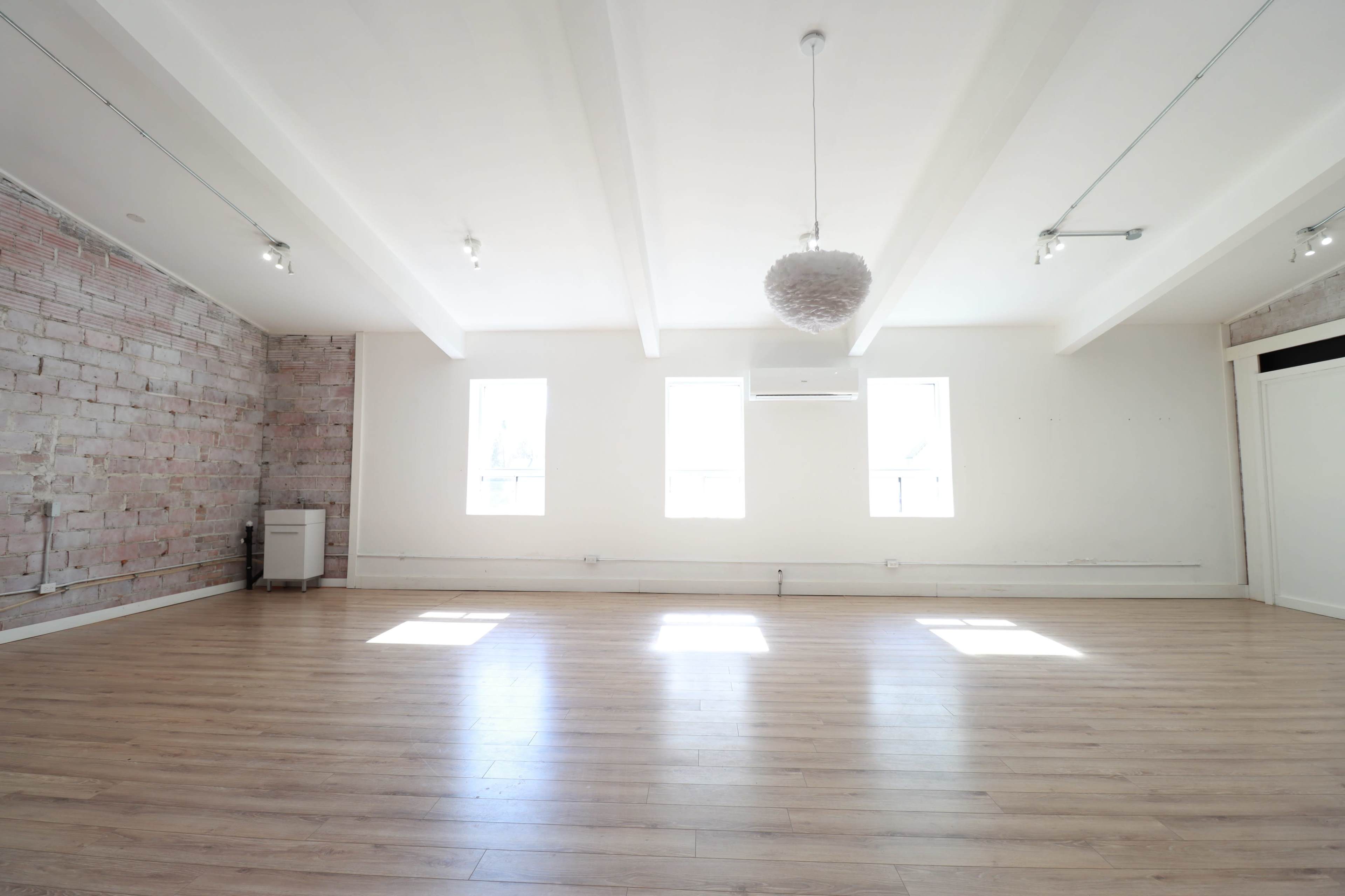 A spacious, well-lit room features wooden flooring, exposed brick walls, and windows allowing natural light to enter.