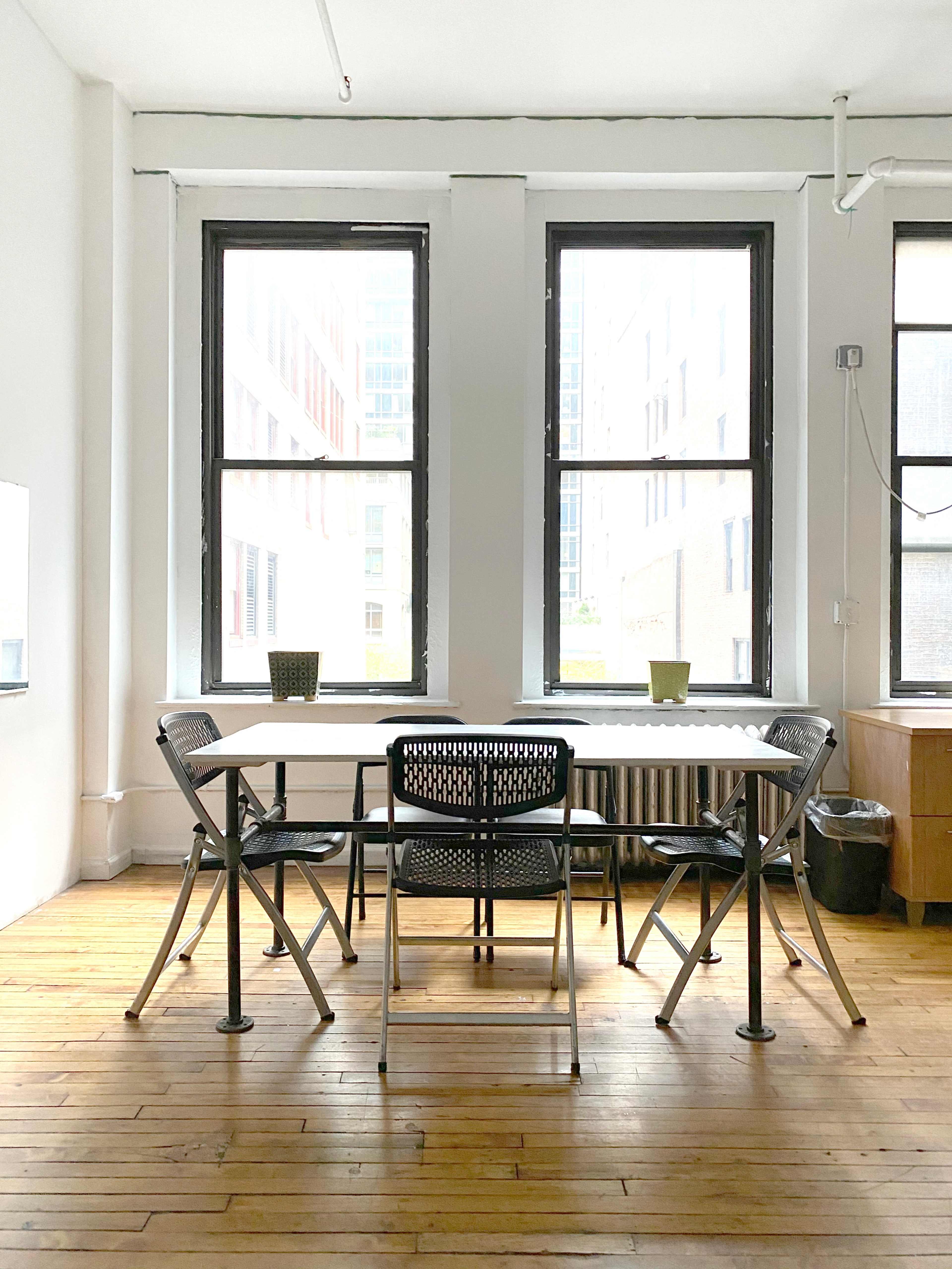 A simple dining table surrounded by four chairs is positioned between two large windows in a bright, minimalist room with wooden flooring.
