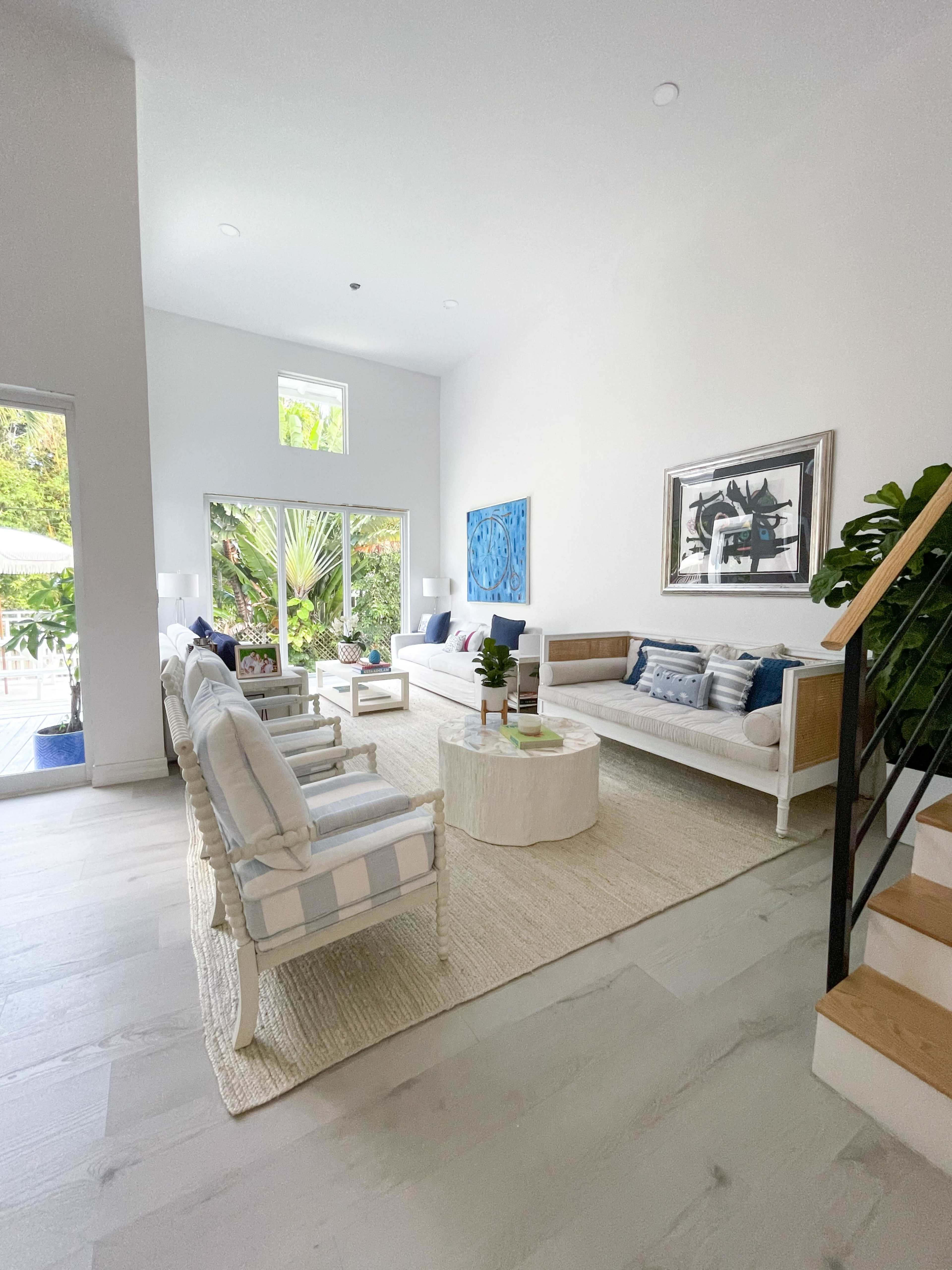 A bright living room with a neutral color palette, featuring two sofas, chairs, and a round coffee table surrounded by large windows and greenery outside.