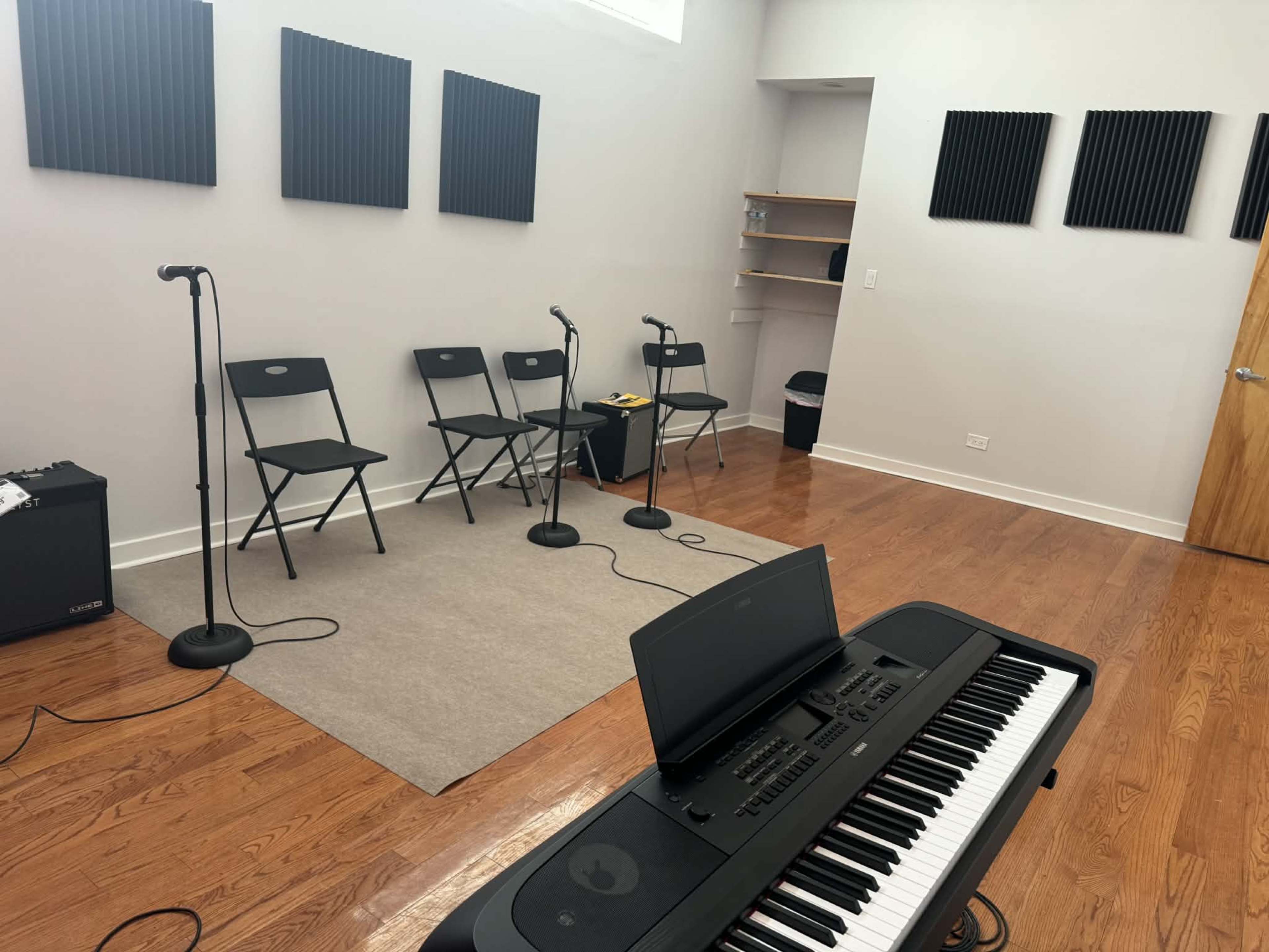 The image shows a soundproof room equipped with a digital piano, four microphones on stands, and several folding chairs arranged on a wooden floor.