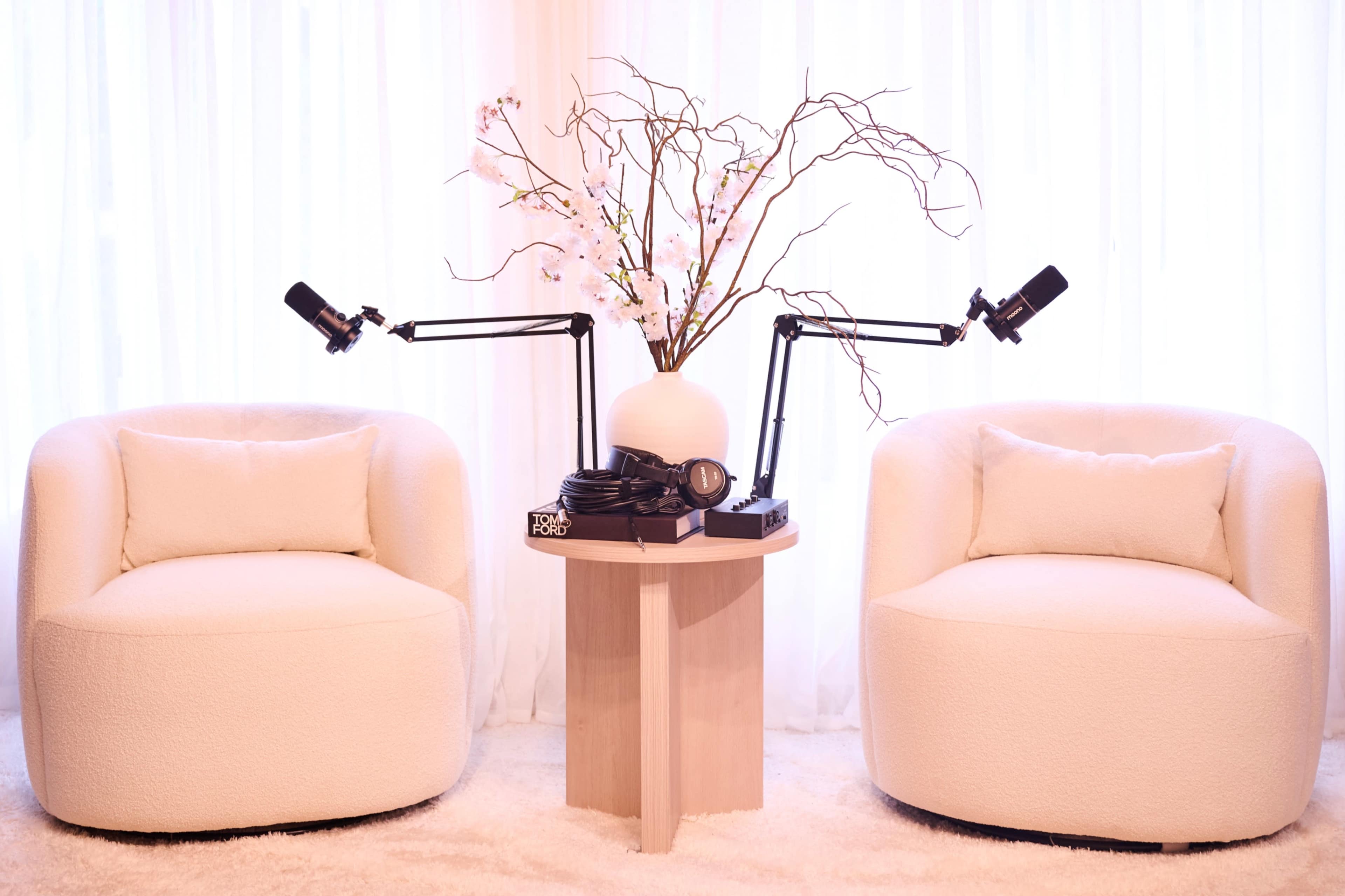 The image features two white, round chairs positioned on either side of a small wooden table with a decorative vase of flowers and two microphones mounted on adjustable arms.