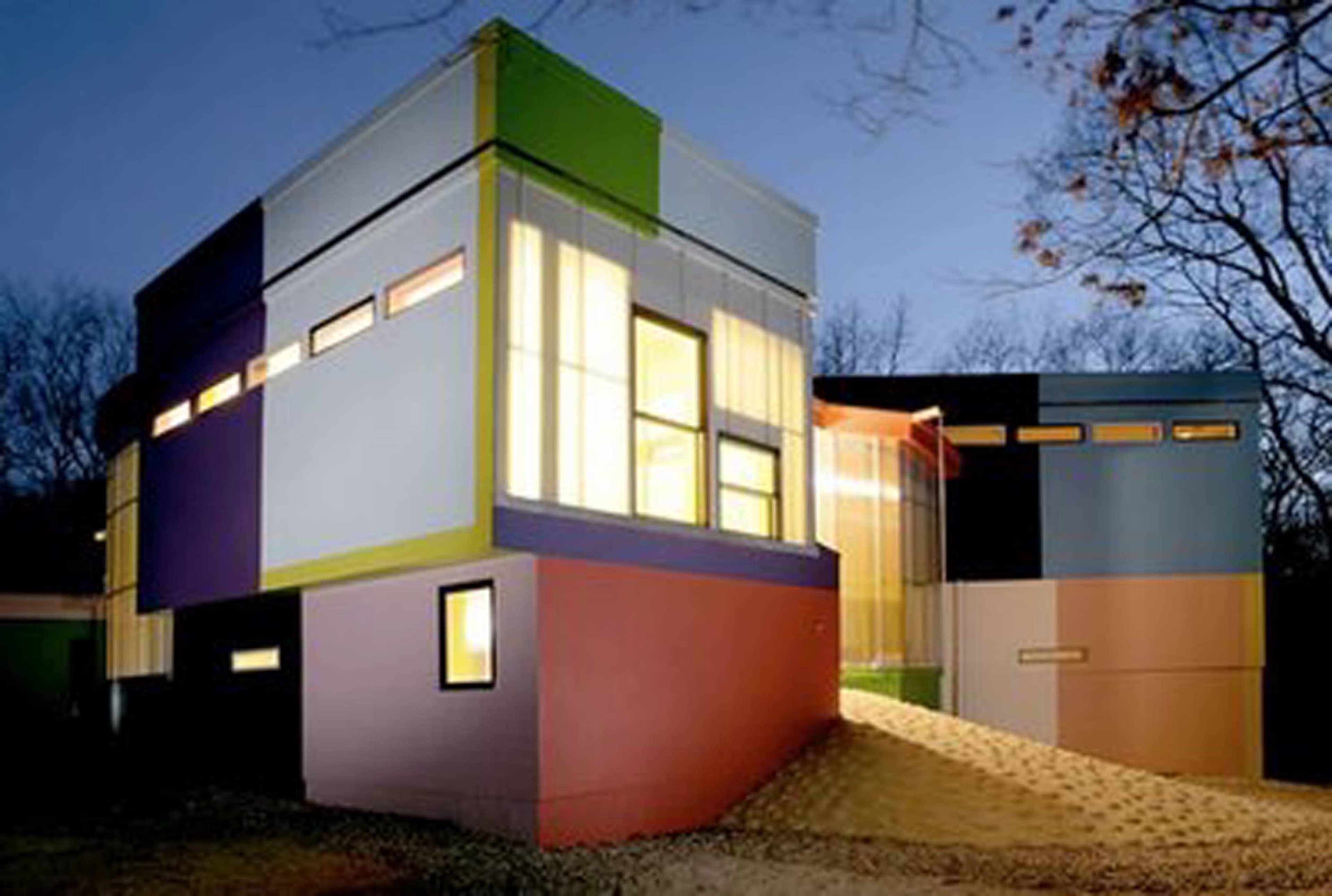 A modern, multicolored house is illuminated at dusk, showcasing a mix of angular shapes and large windows.