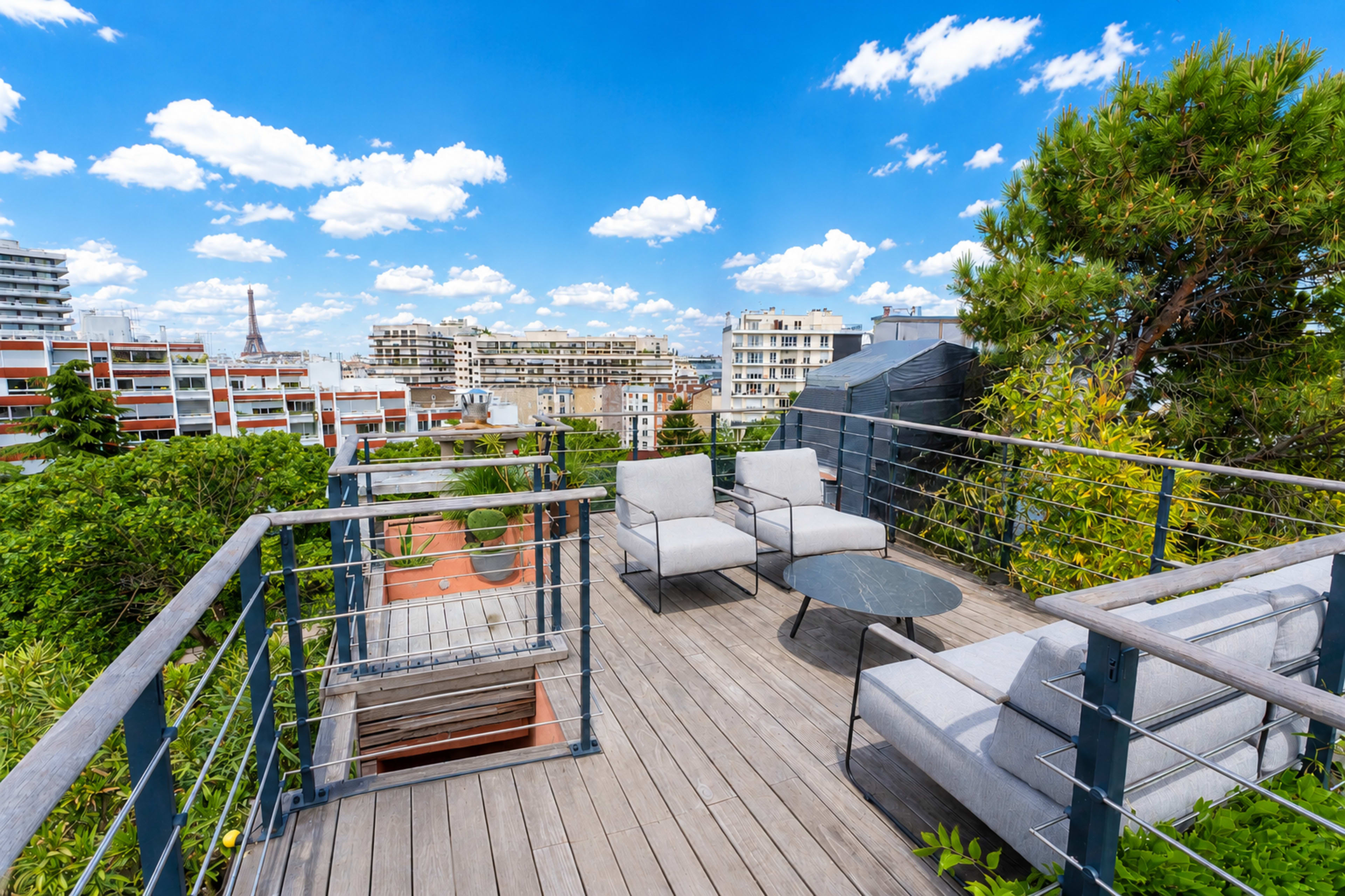 The image shows a rooftop terrace with modern furniture, including two chairs and a table, surrounded by greenery and city buildings under a blue sky with clouds.