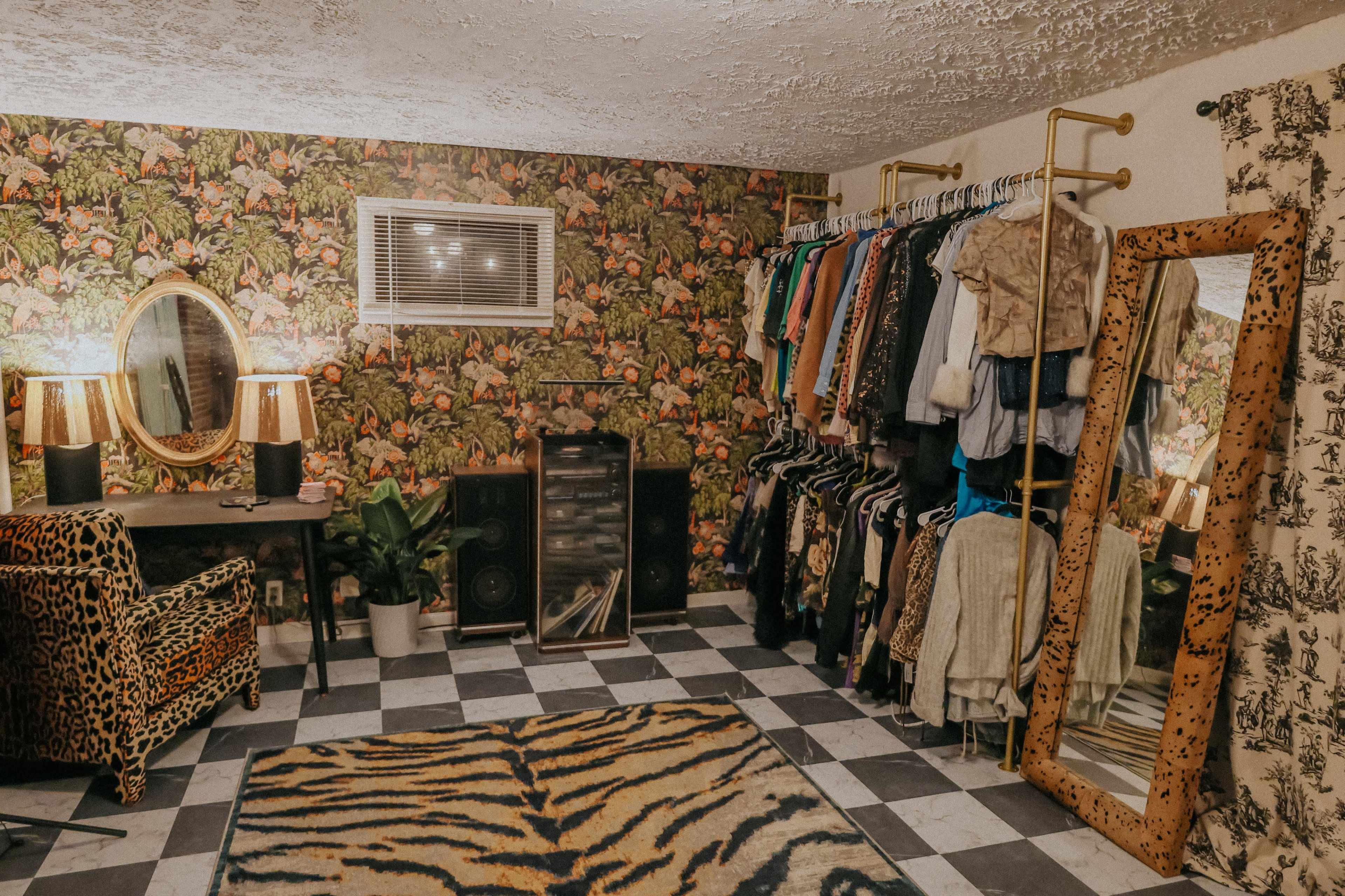 The image shows a vintage-inspired room with floral wallpaper, a clothing rack filled with garments, a large mirror, and a patterned rug on the floor.