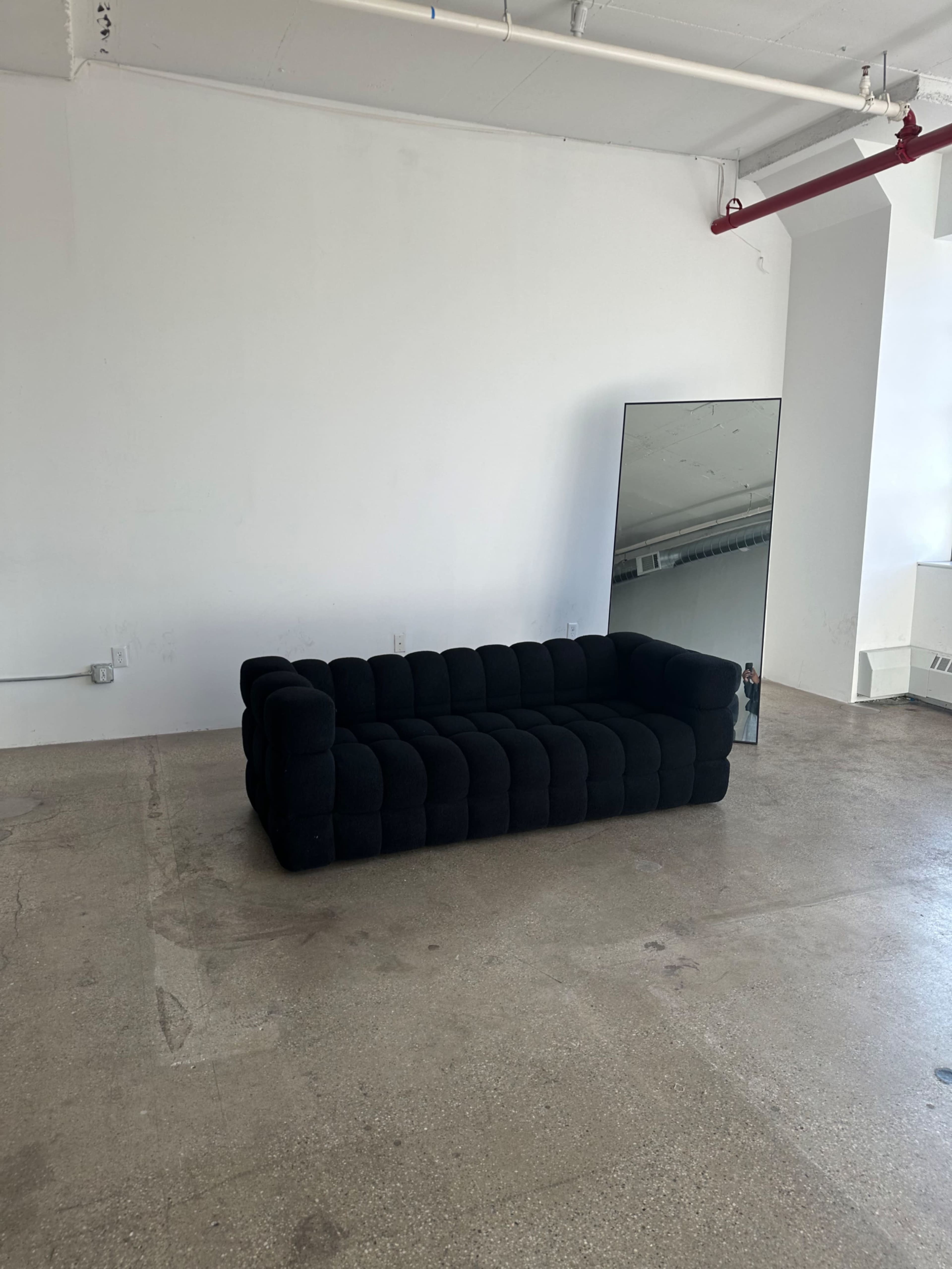 The image shows a black, tufted sofa in an empty room with exposed concrete flooring and a large mirror leaning against the wall.