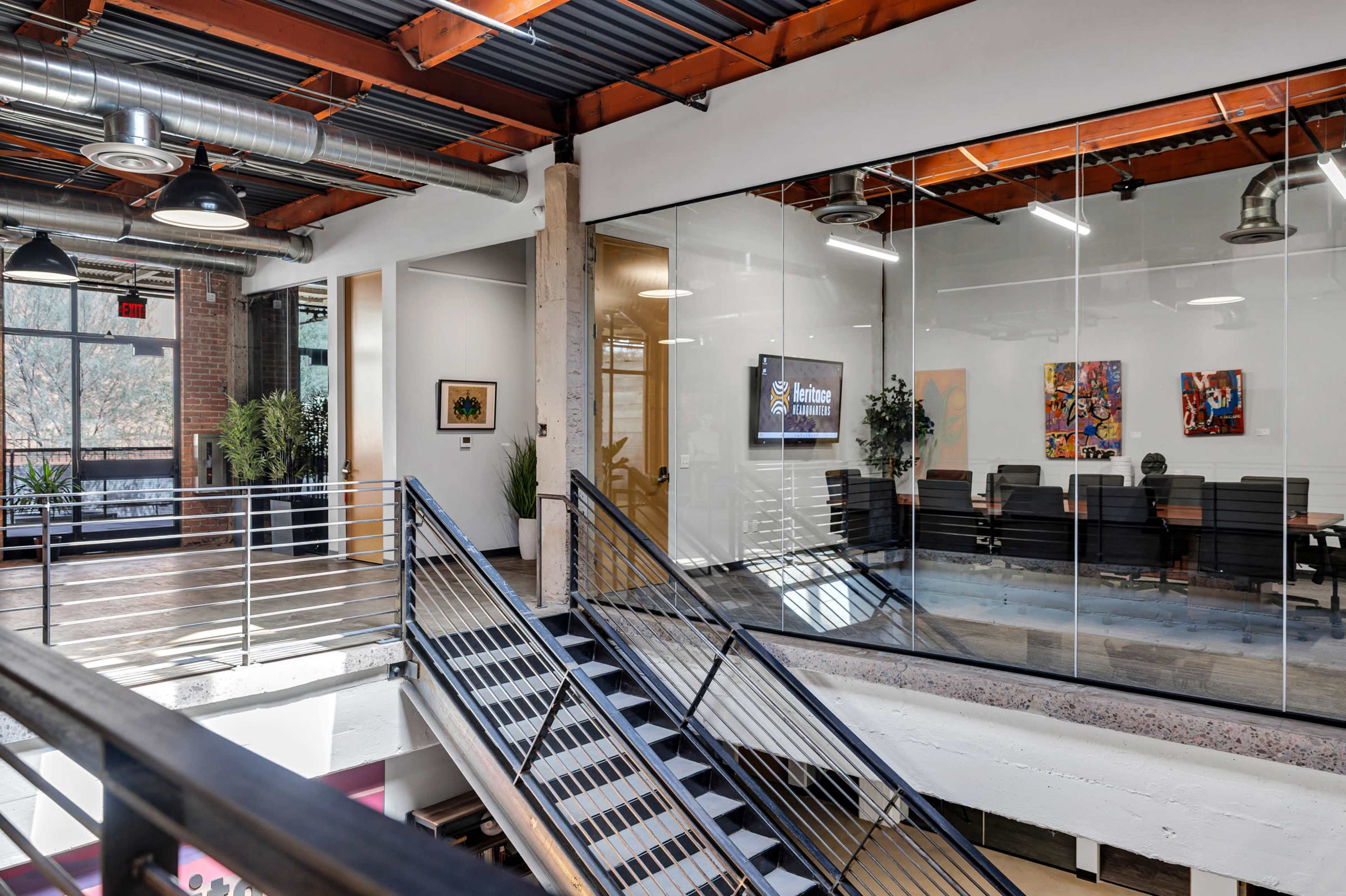 The image shows a modern office space with glass walls, a staircase leading to an upper level, and various artworks displayed on the walls.