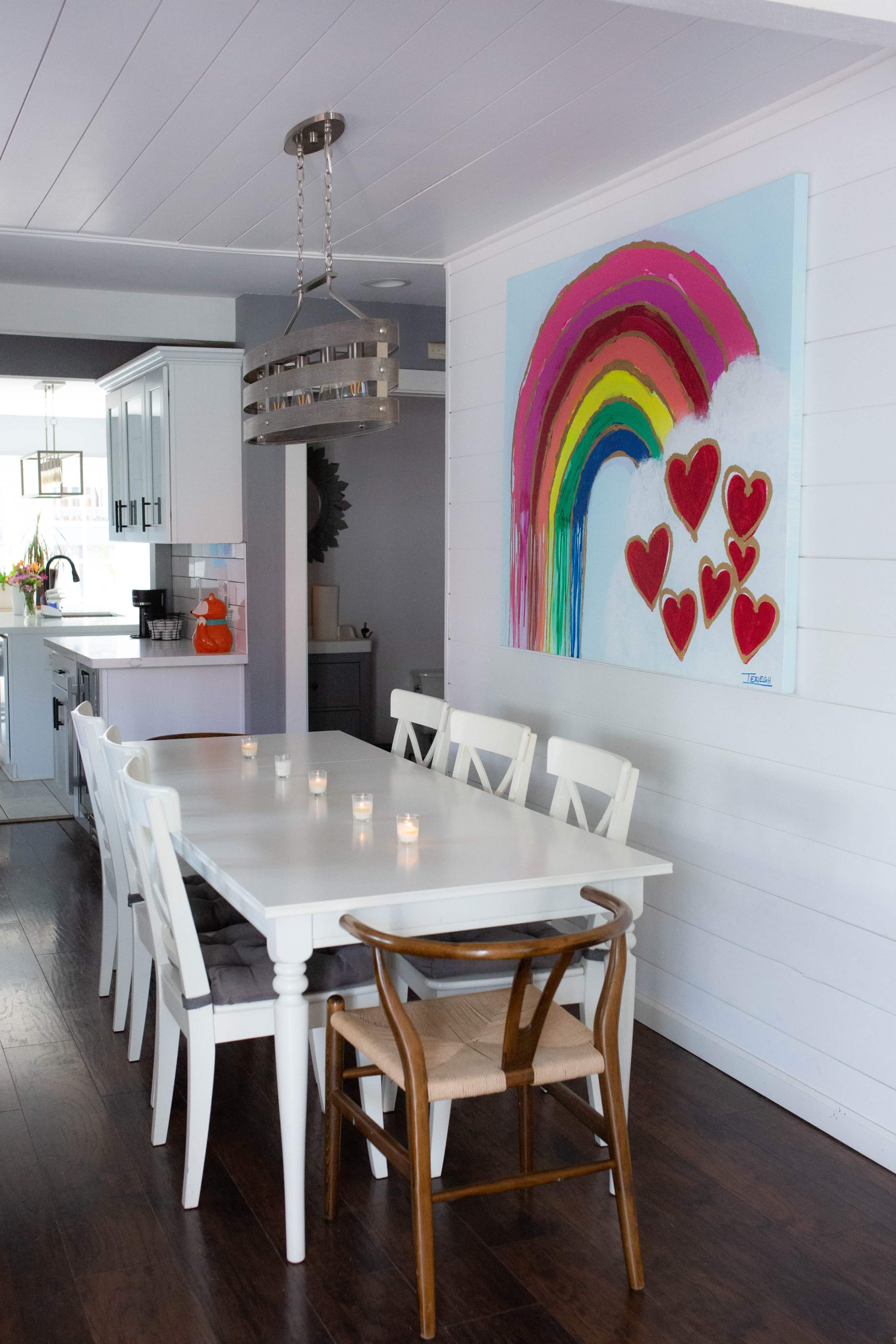 A dining area features a long white table surrounded by chairs, with a colorful rainbow and heart artwork hanging on the wall.