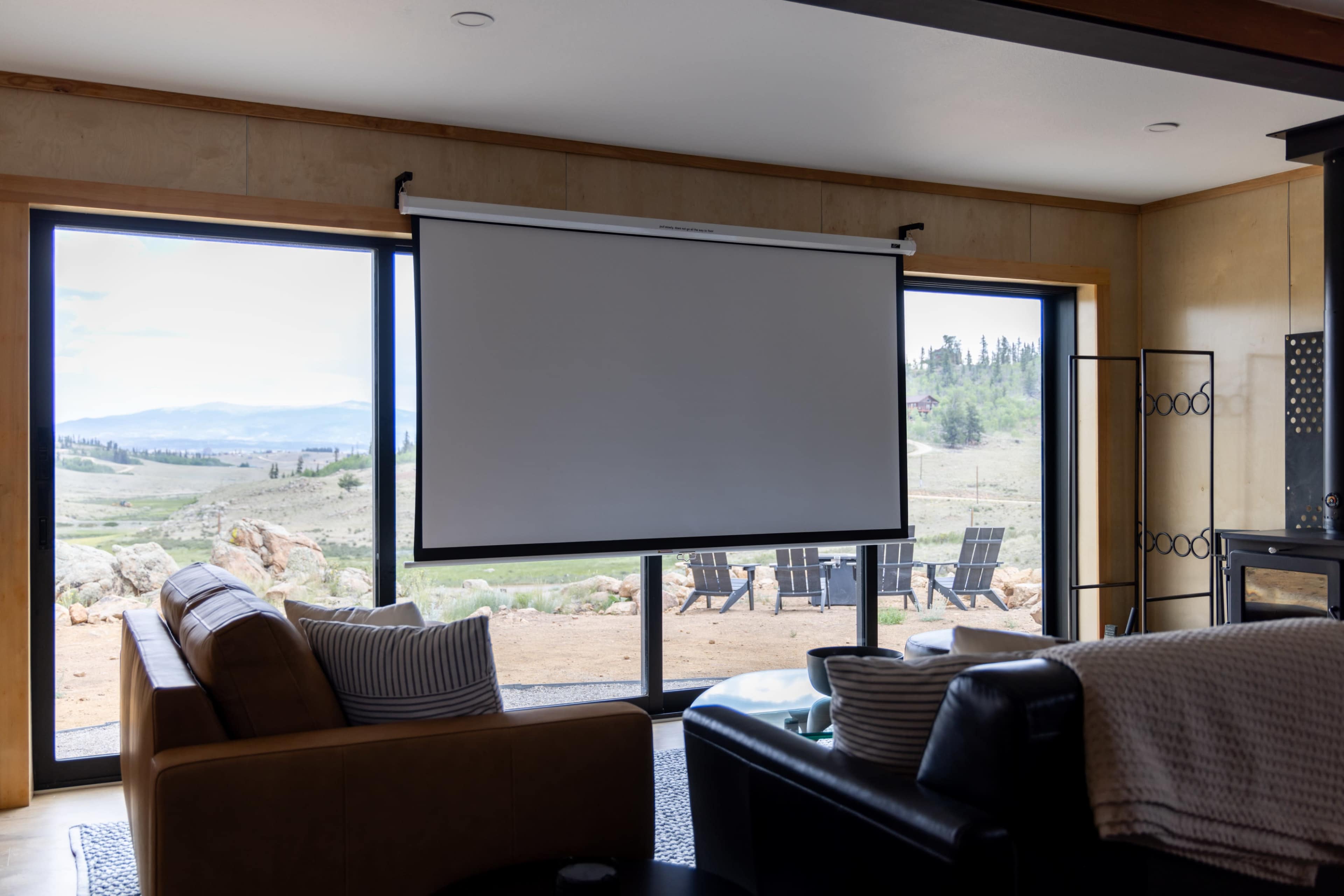 A large, blank projection screen hangs between two floor-to-ceiling windows, showcasing a view of a landscaped outdoor area with seating.