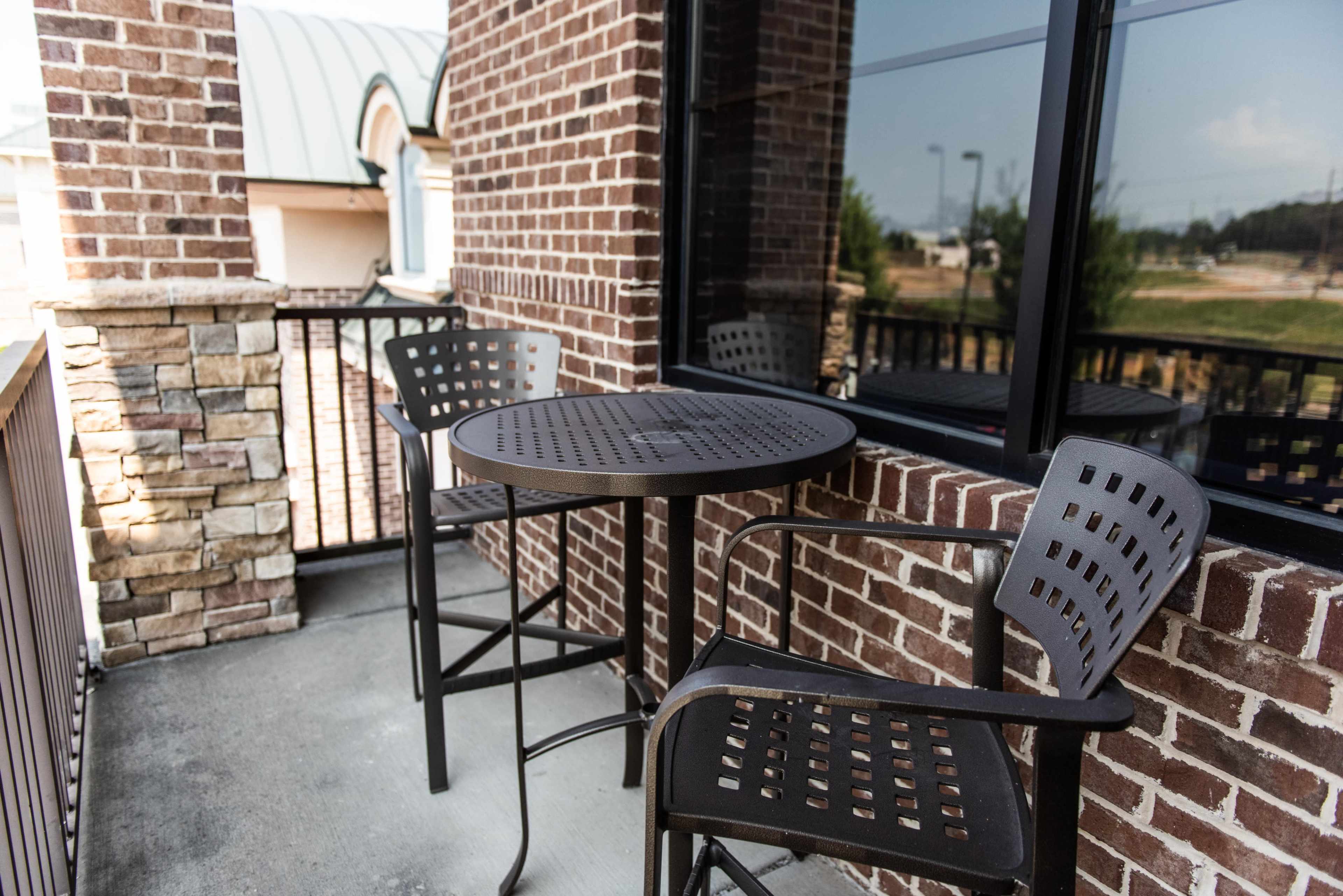 A small balcony features a metal table and two chairs made of perforated black material, set against a brick wall and a large window.
