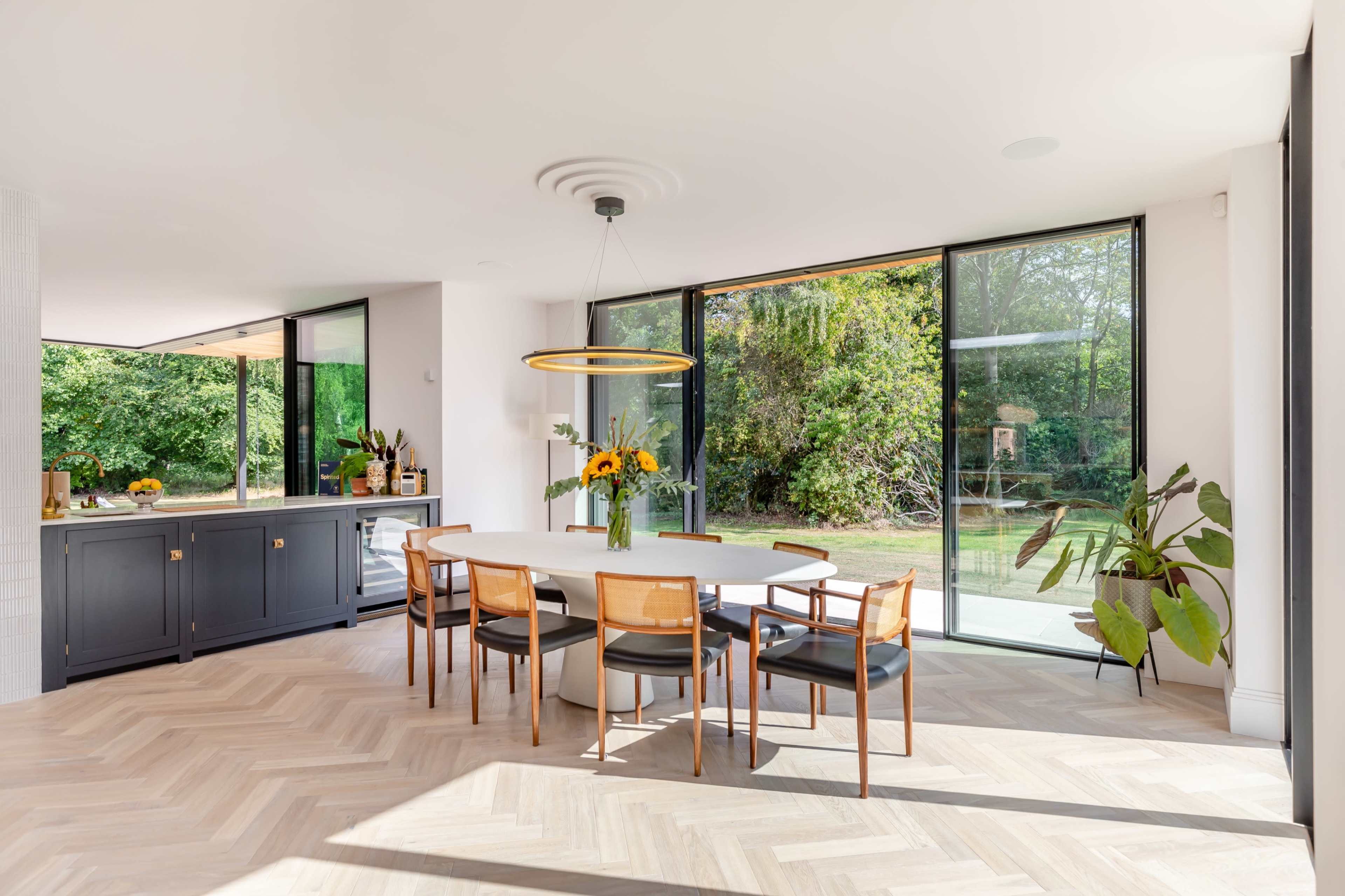 The image shows a modern dining area with a round white table surrounded by six chairs, large windows showcasing a garden view, and a side cabinet with decorative plants.