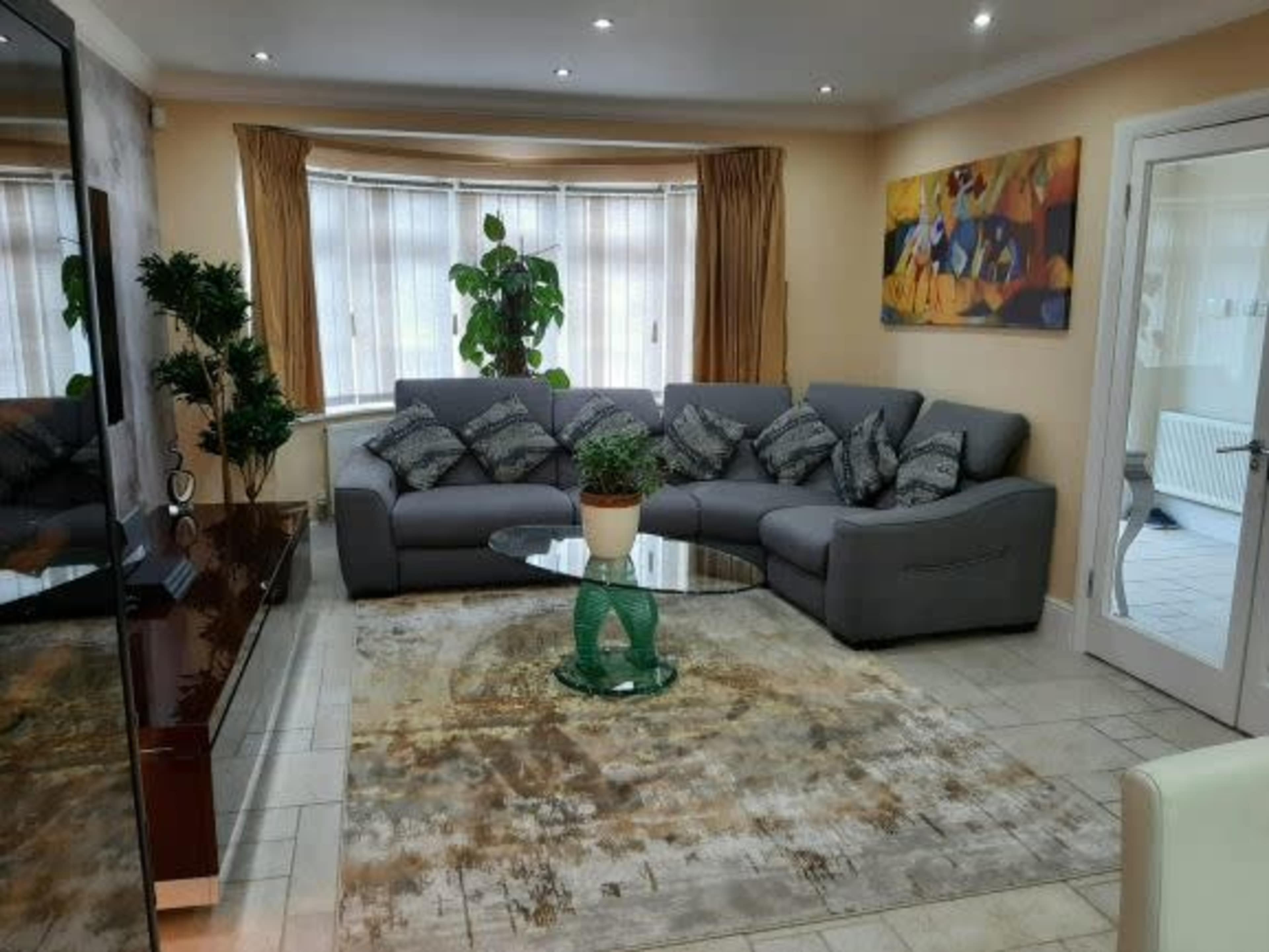 The living room features a gray sectional sofa with decorative cushions, a glass coffee table on a patterned rug, and a large window with indoor plants.