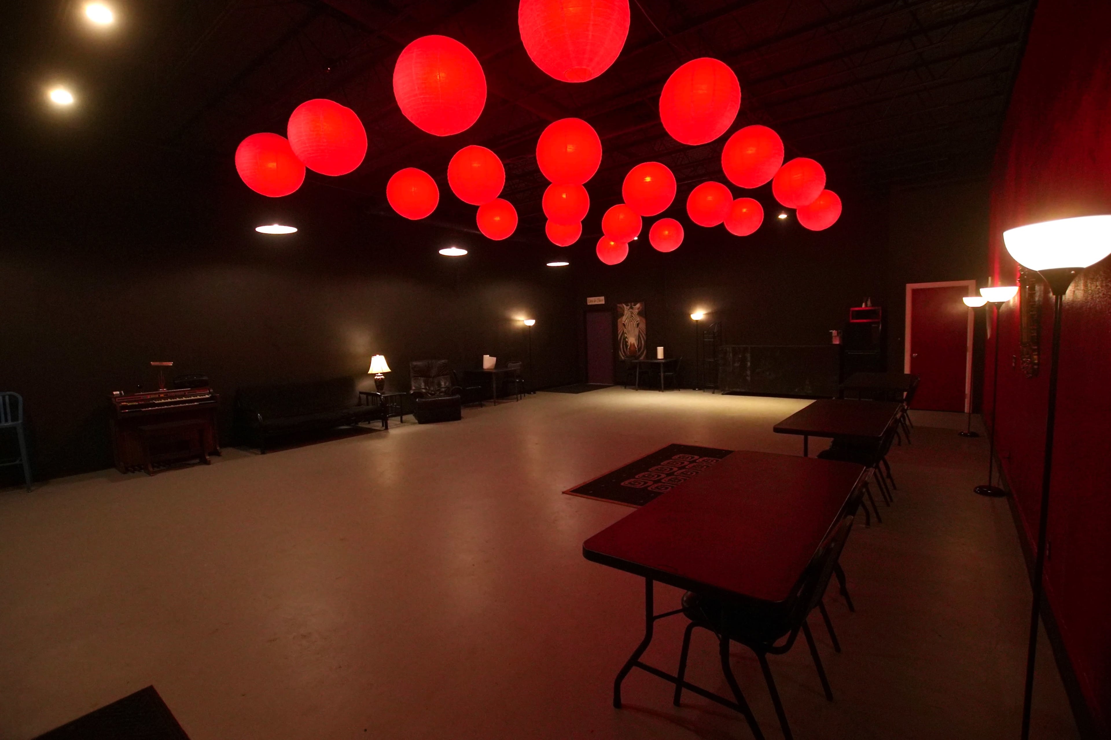 A spacious room with a gray floor, dark walls, and several red paper lanterns hanging from the ceiling, accompanied by tables and couches along the sides.