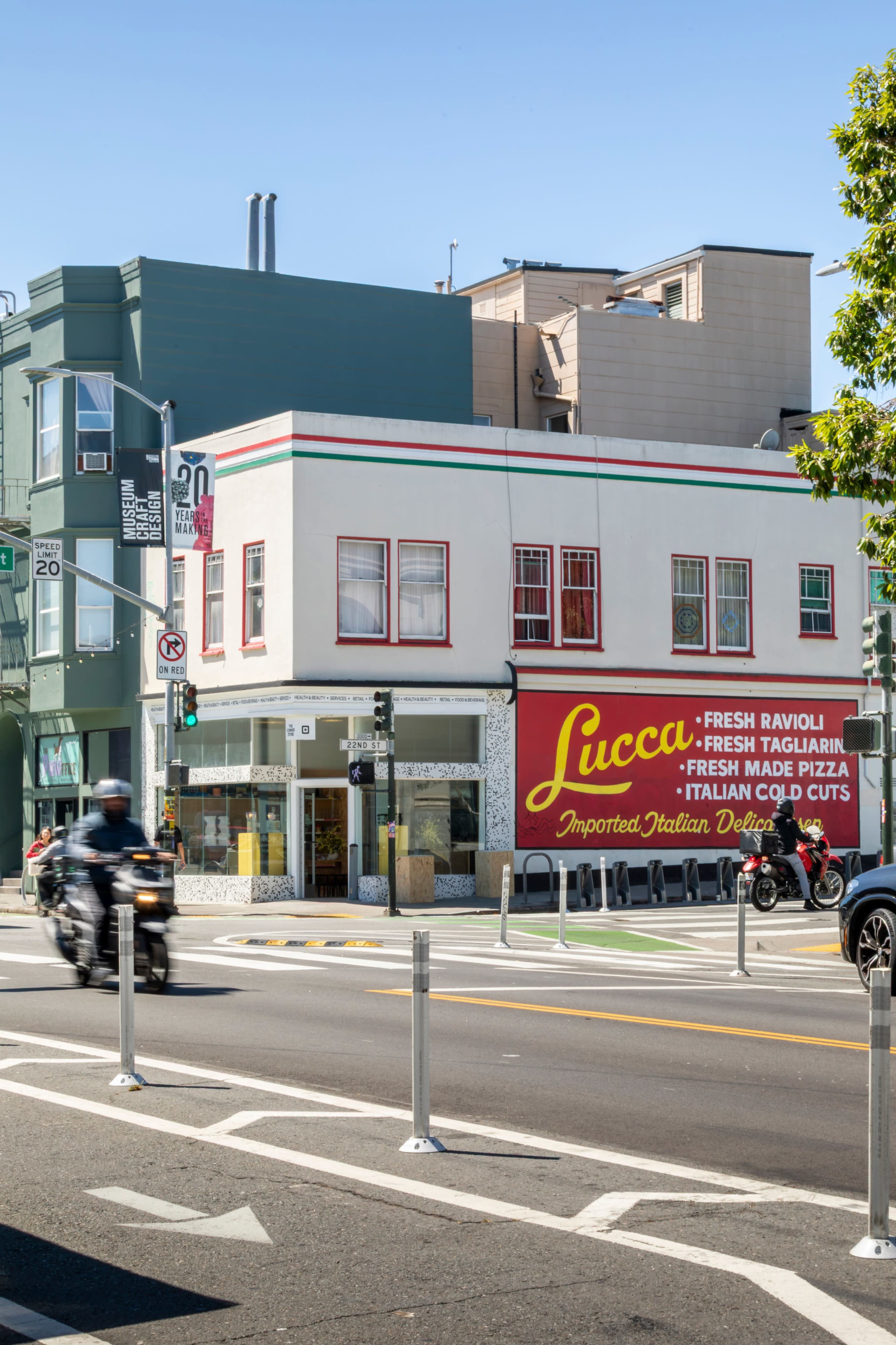 A corner building features a vibrant store sign for Lucca, advertising fresh Italian food, with vehicles passing by on the street.