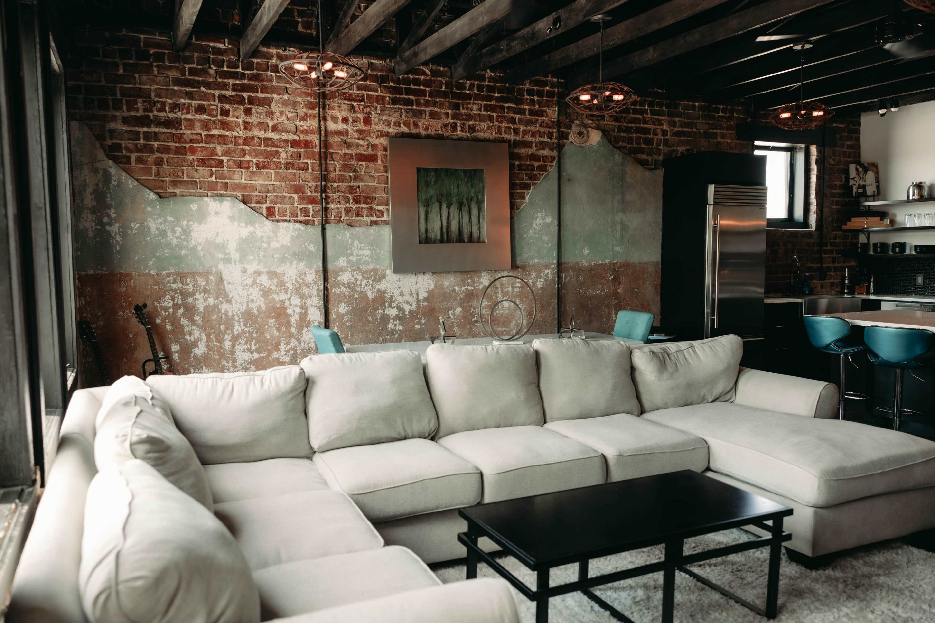 The image shows a modern living room with a large sectional sofa, a black coffee table, exposed brick walls, and pendant lighting.