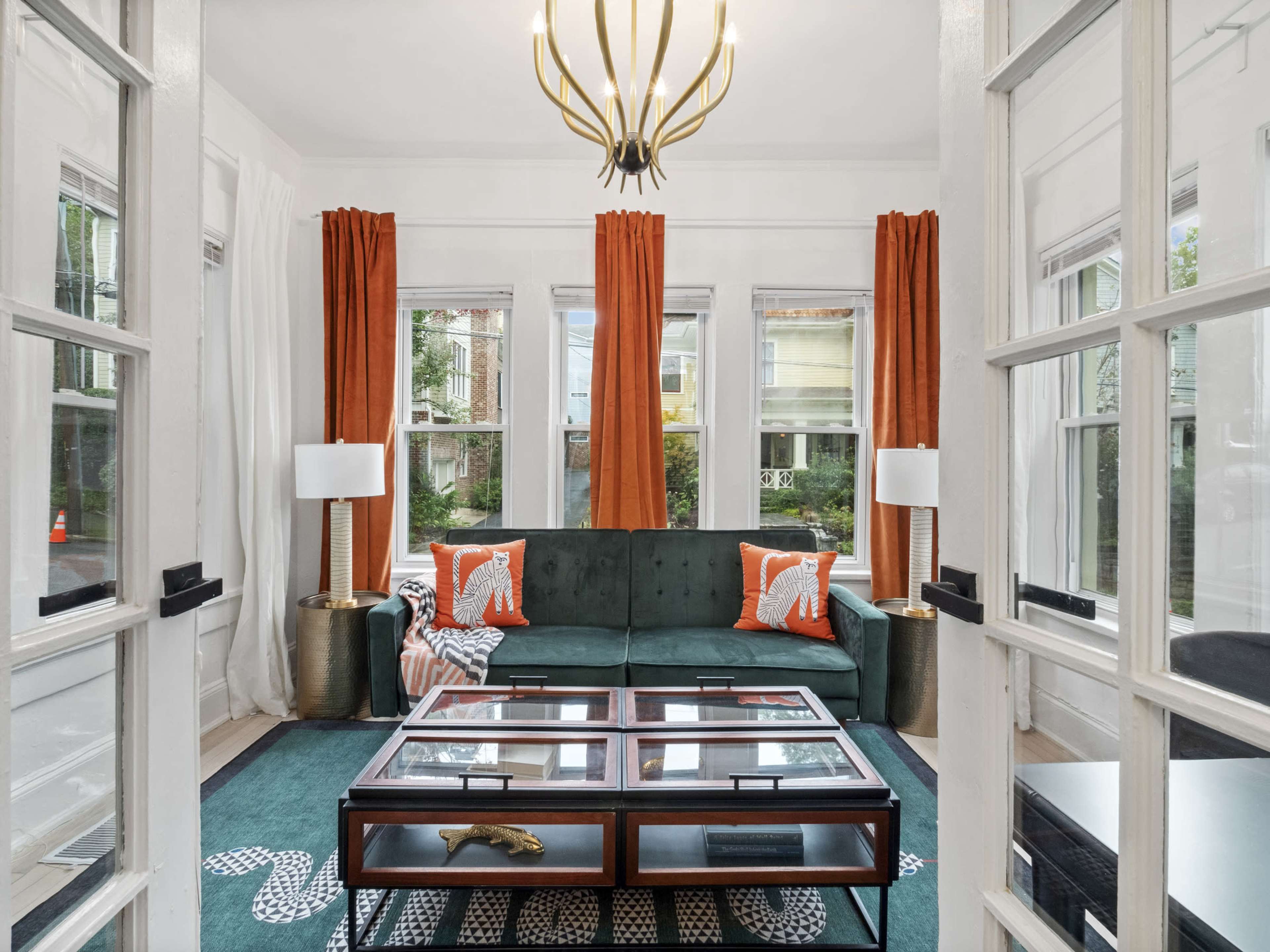 The image shows a bright living room featuring a green sofa, large windows with orange curtains, and a coffee table with a glass top.