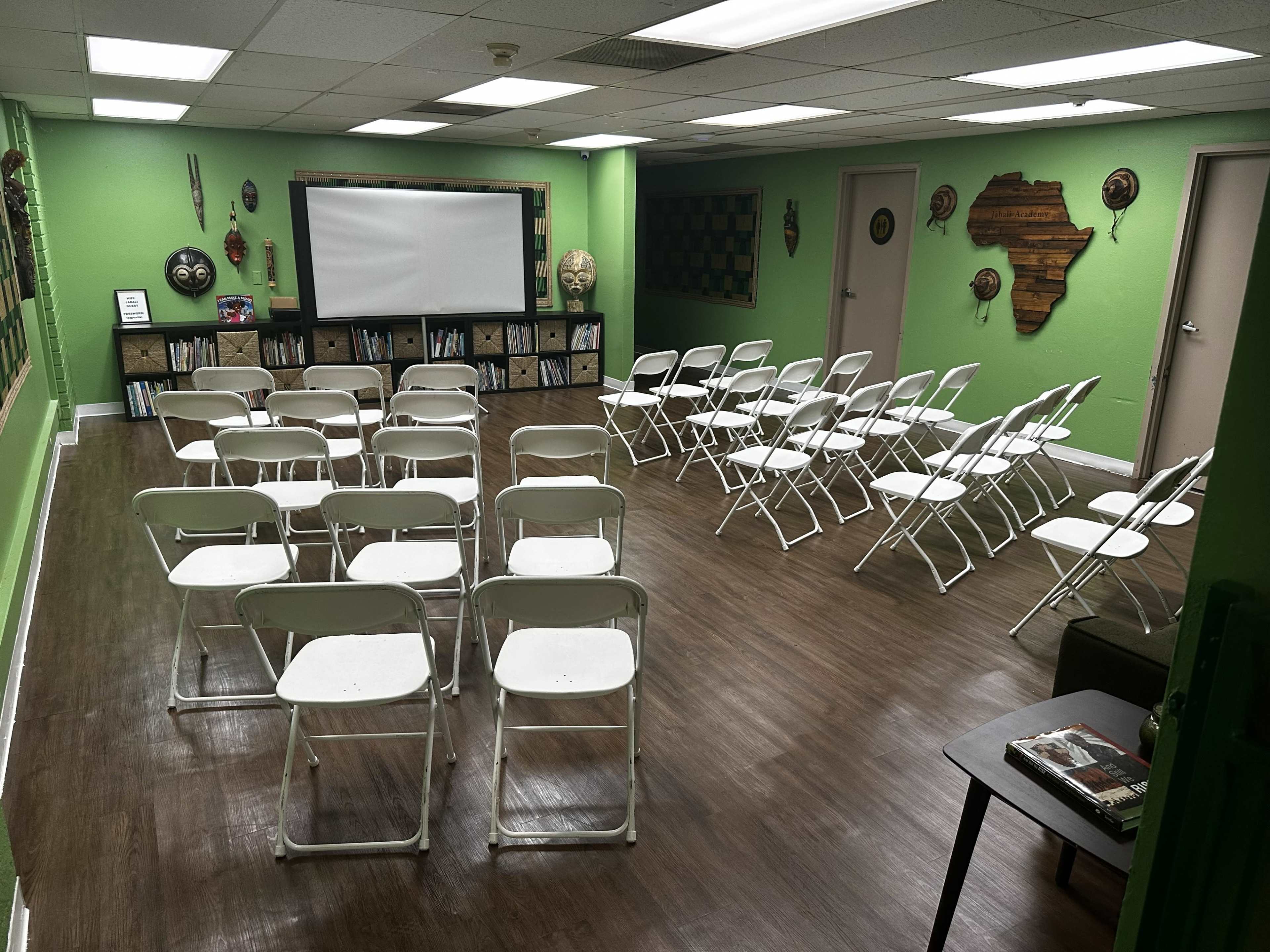The image shows a classroom-like setting with rows of white folding chairs arranged in front of a blank screen on a green wall, with decorative elements and shelves visible in the background.