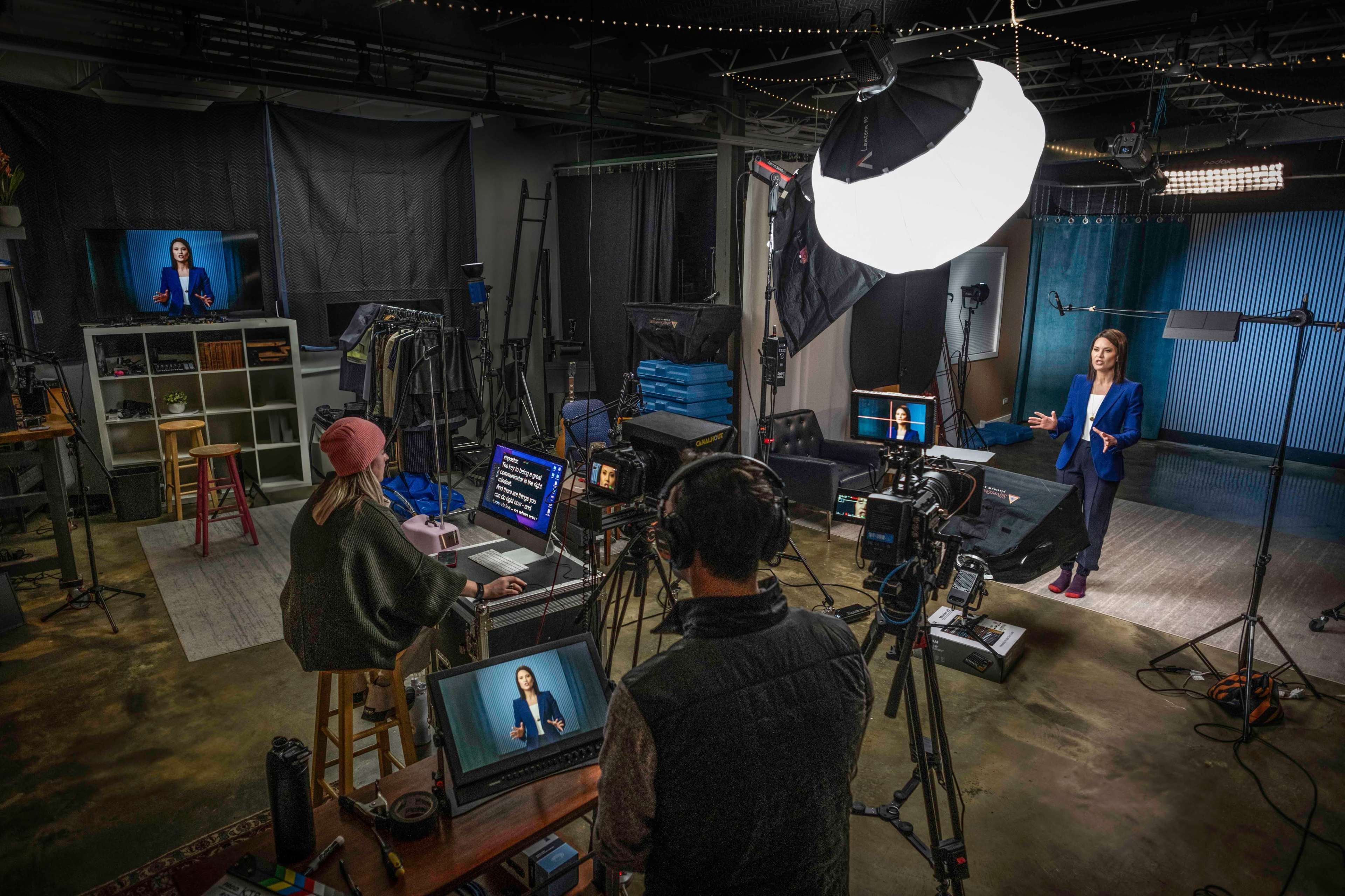 A studio setup features a woman speaking on a video screen while two individuals work at computers and cameras in the foreground.