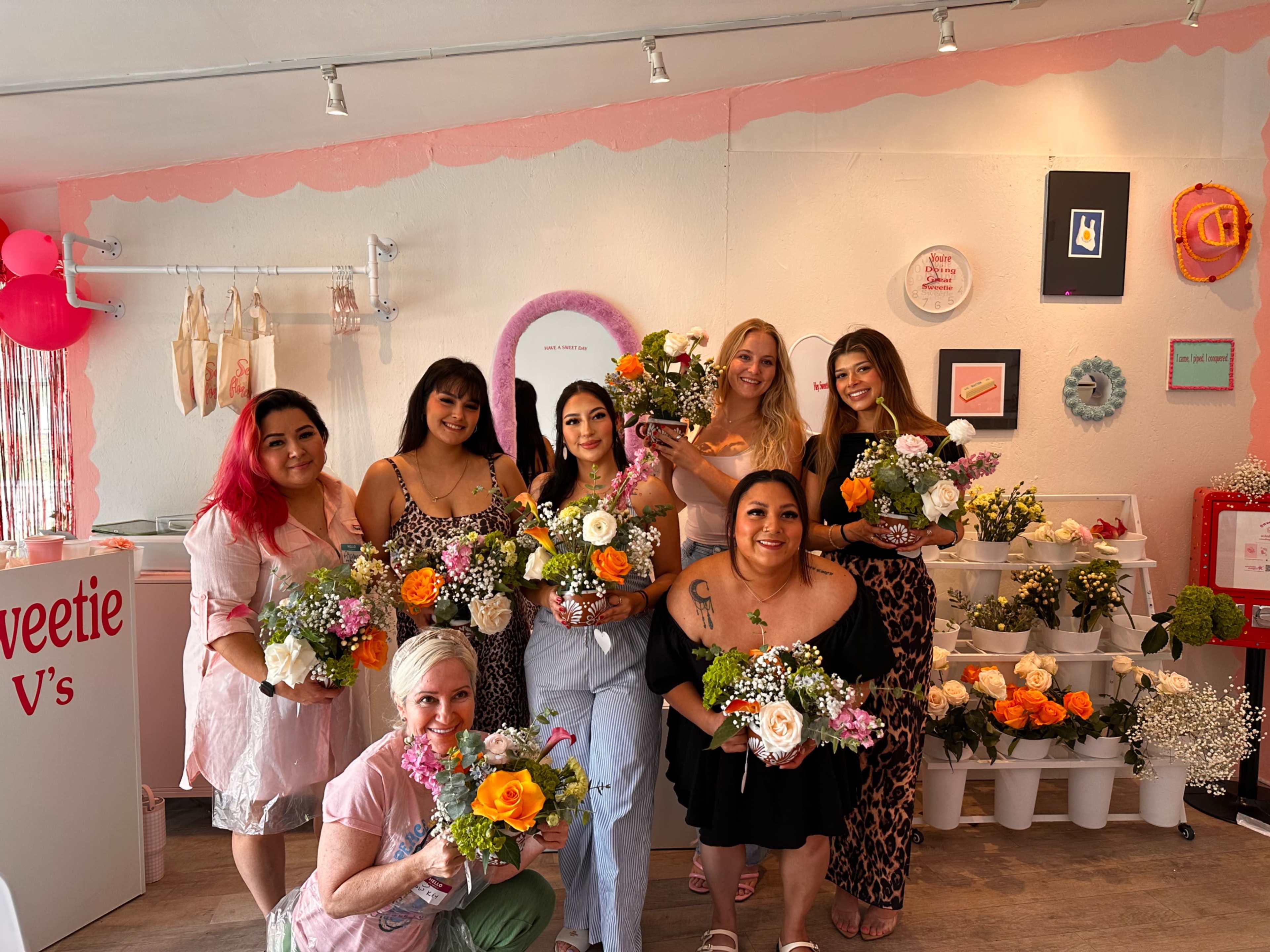 A group of eight women stands together in a flower shop, each holding a bouquet of colorful flowers.