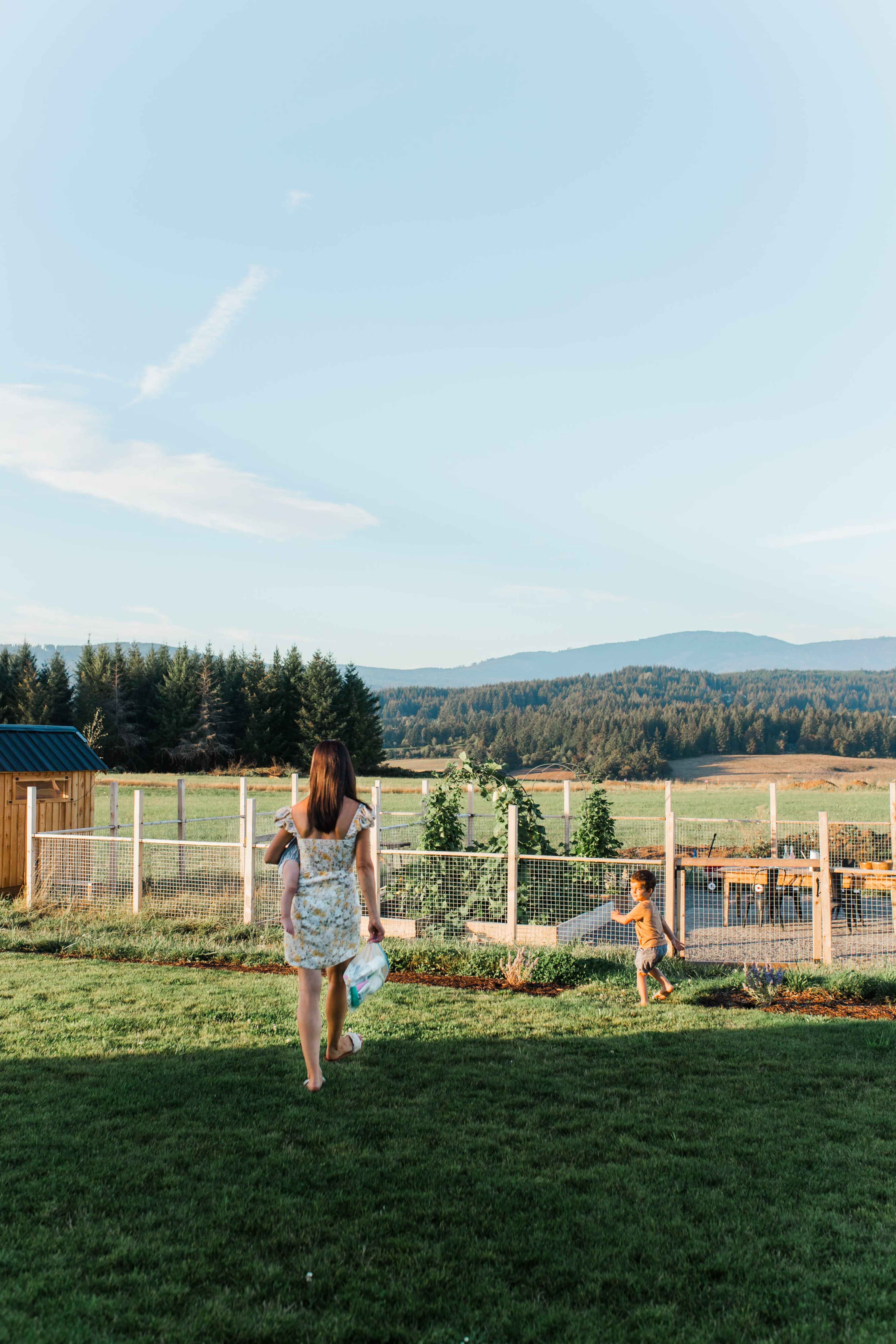 A woman walks through a grassy yard toward a fenced area where a child is playing, with mountains in the background.