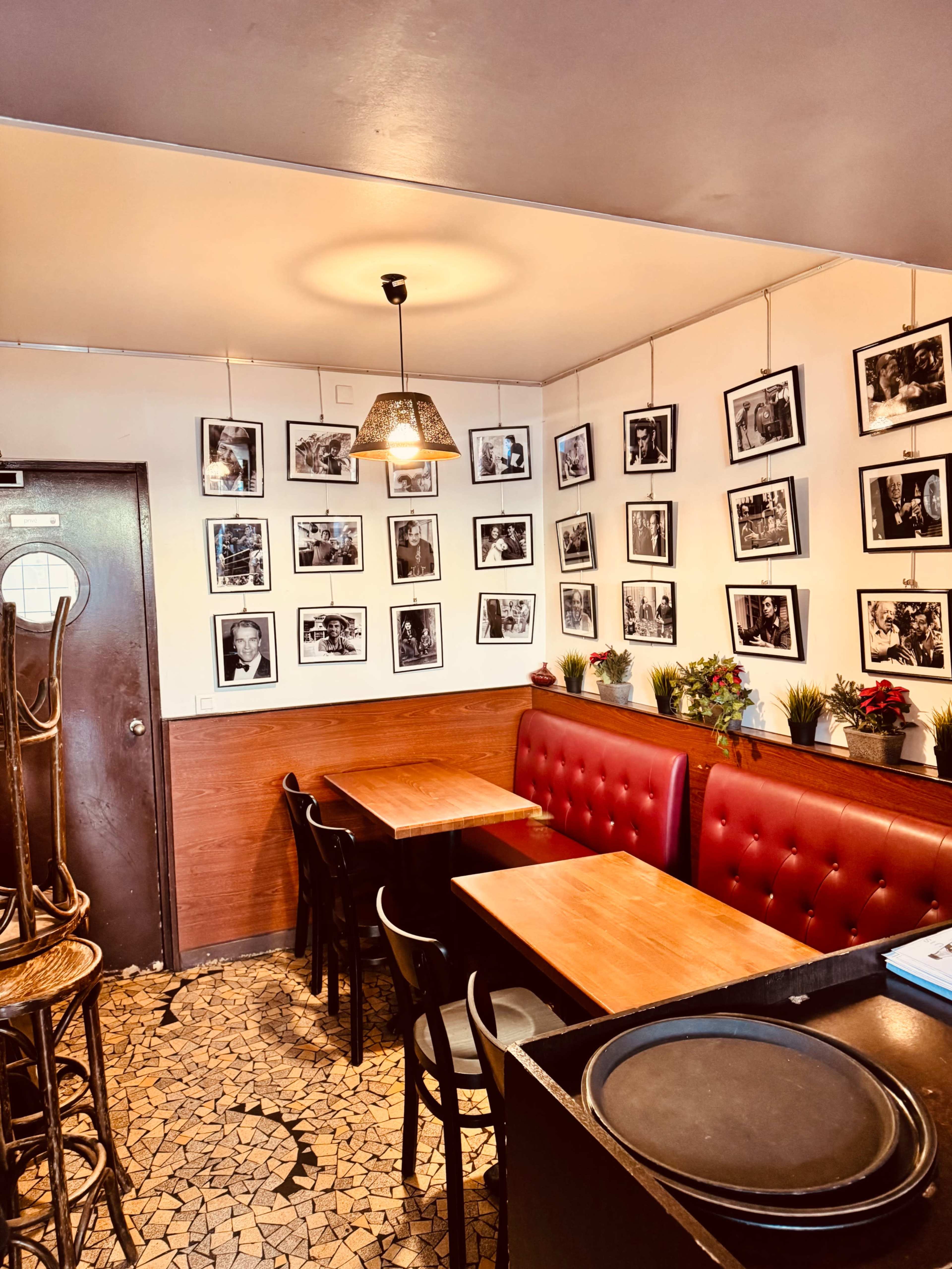The image shows a small café interior featuring two wooden tables, red upholstered seating, and walls adorned with black and white photographs.