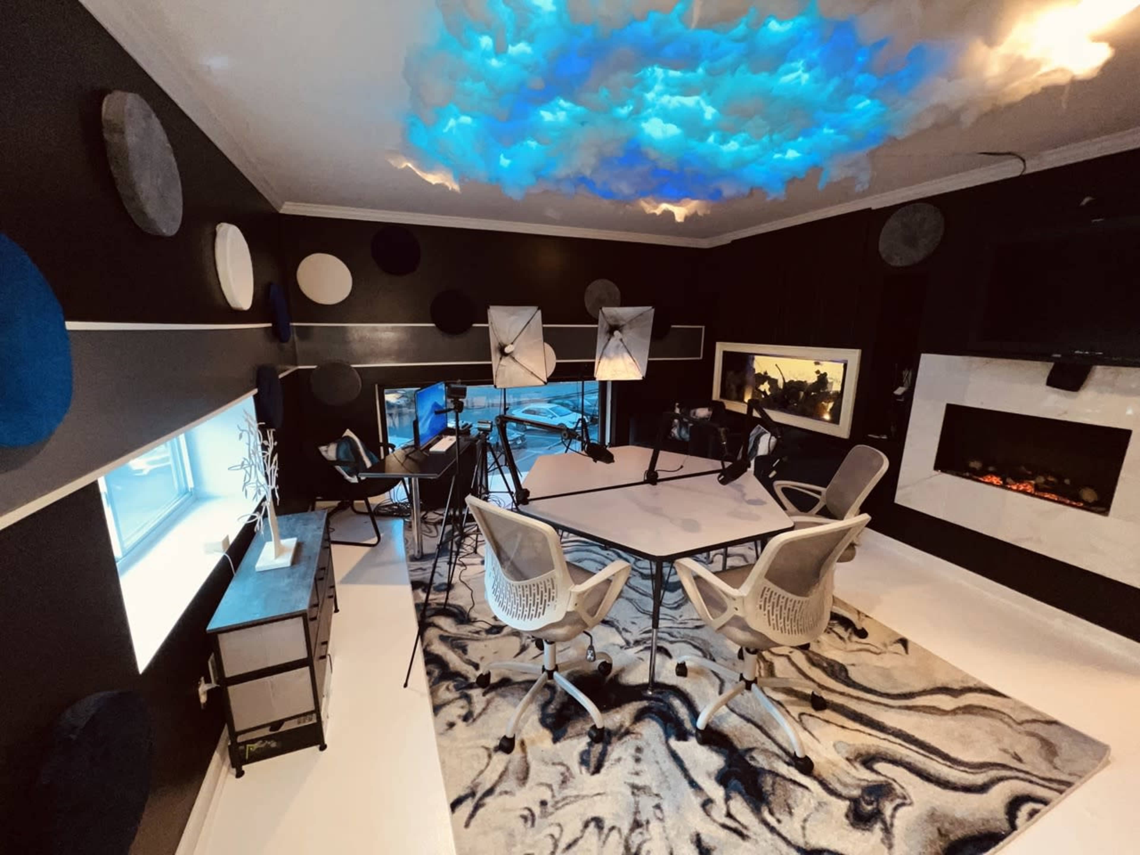 The image shows a modern studio space with a large table, chairs, and lighting equipment, featuring a textured rug and artistic ceiling design.