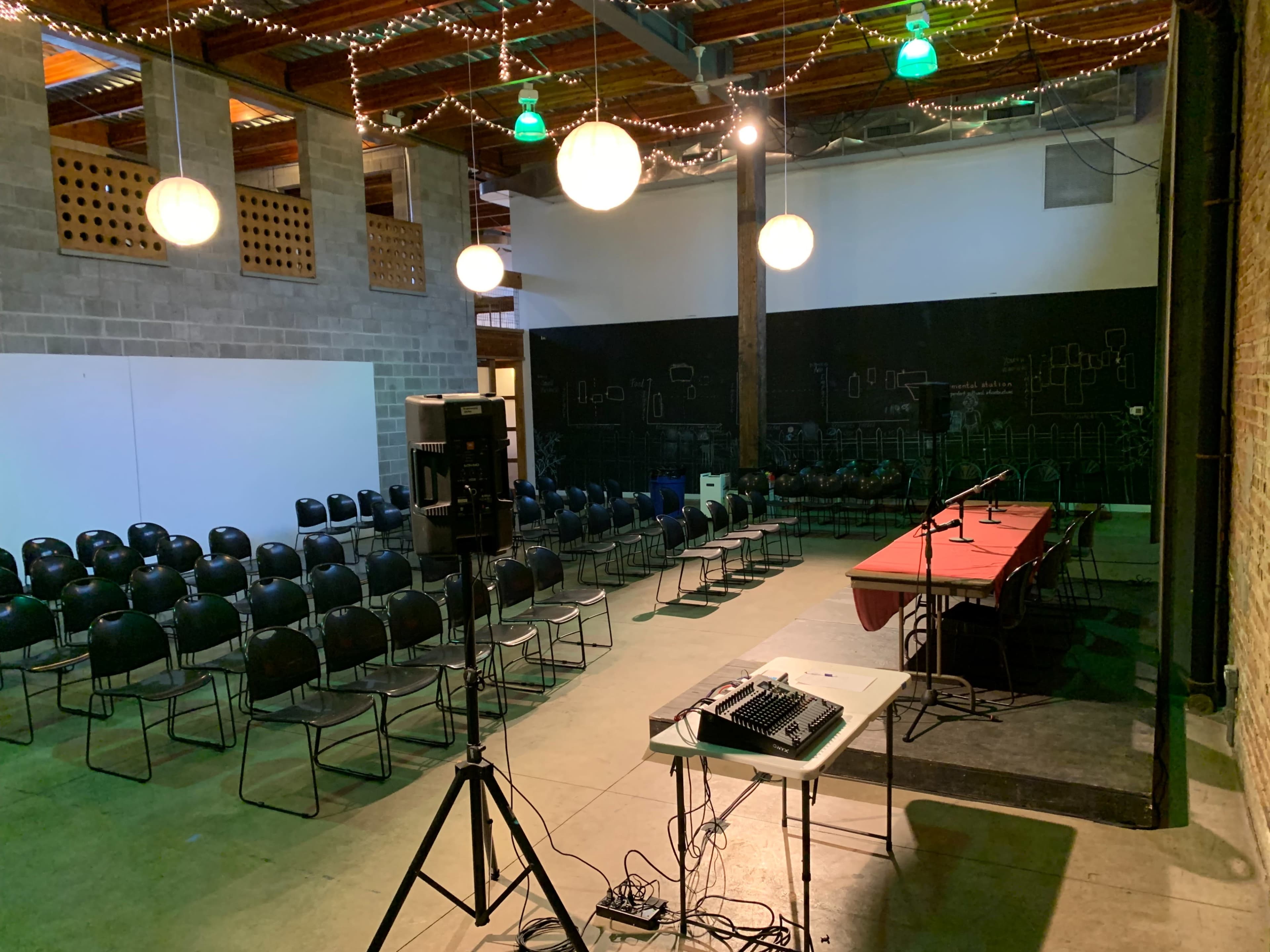 A room with rows of black chairs facing a stage area, illuminated by hanging lights and featuring a table with audio equipment.