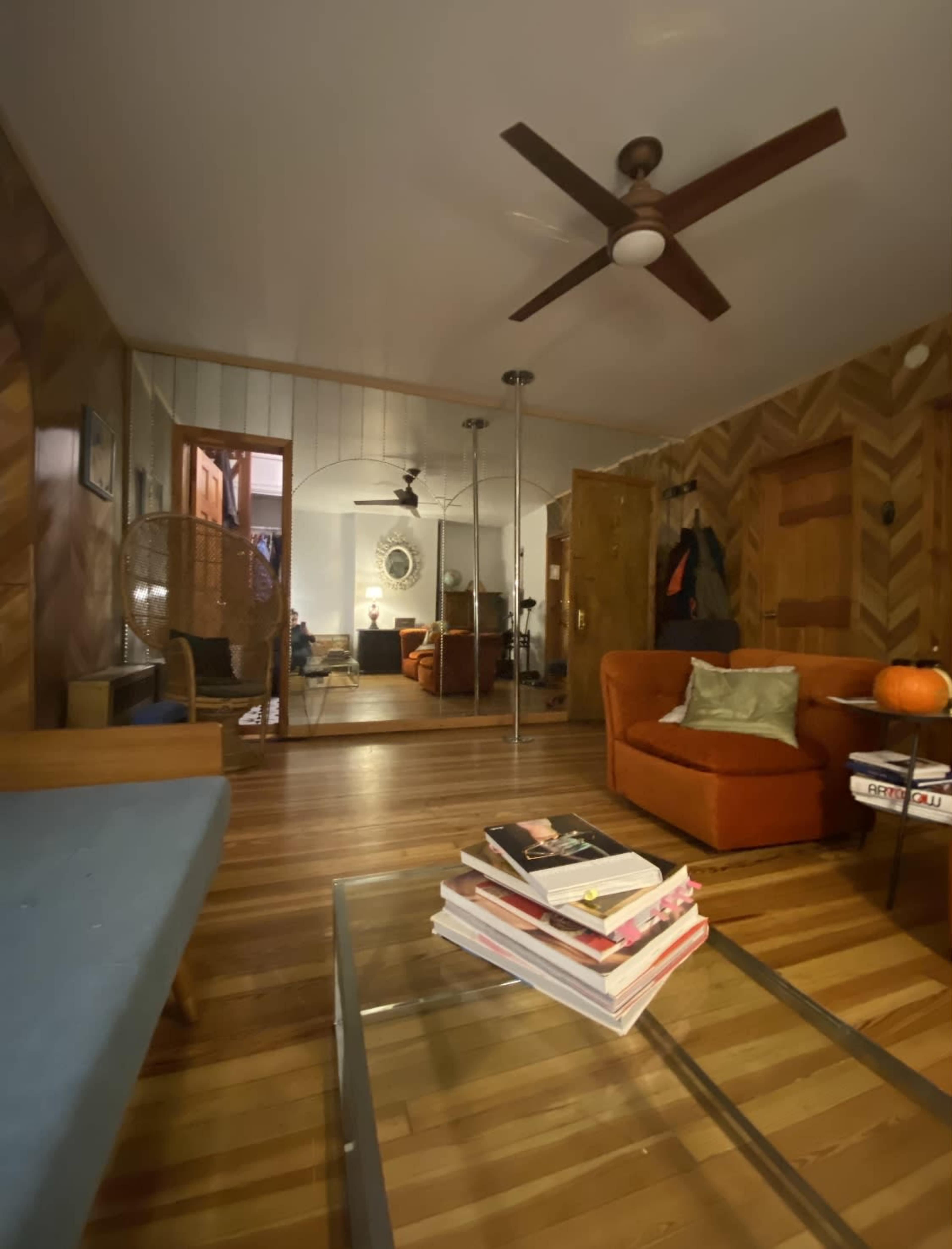 A cozy living room features a wooden floor, a blue sofa, an orange armchair, and a coffee table stacked with books.