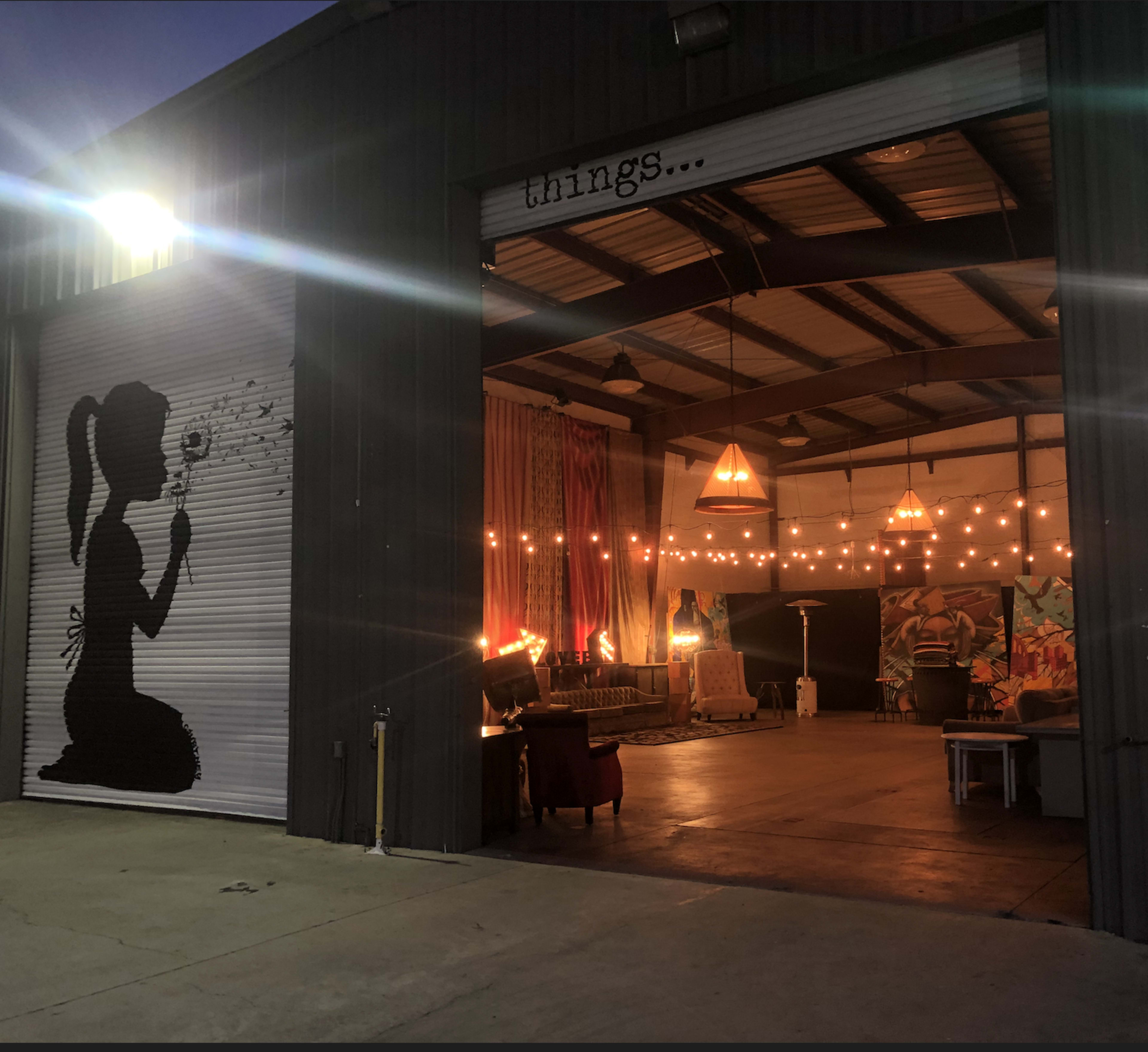 A large industrial garage door is open, revealing an indoor space decorated with lights, art, and furniture.