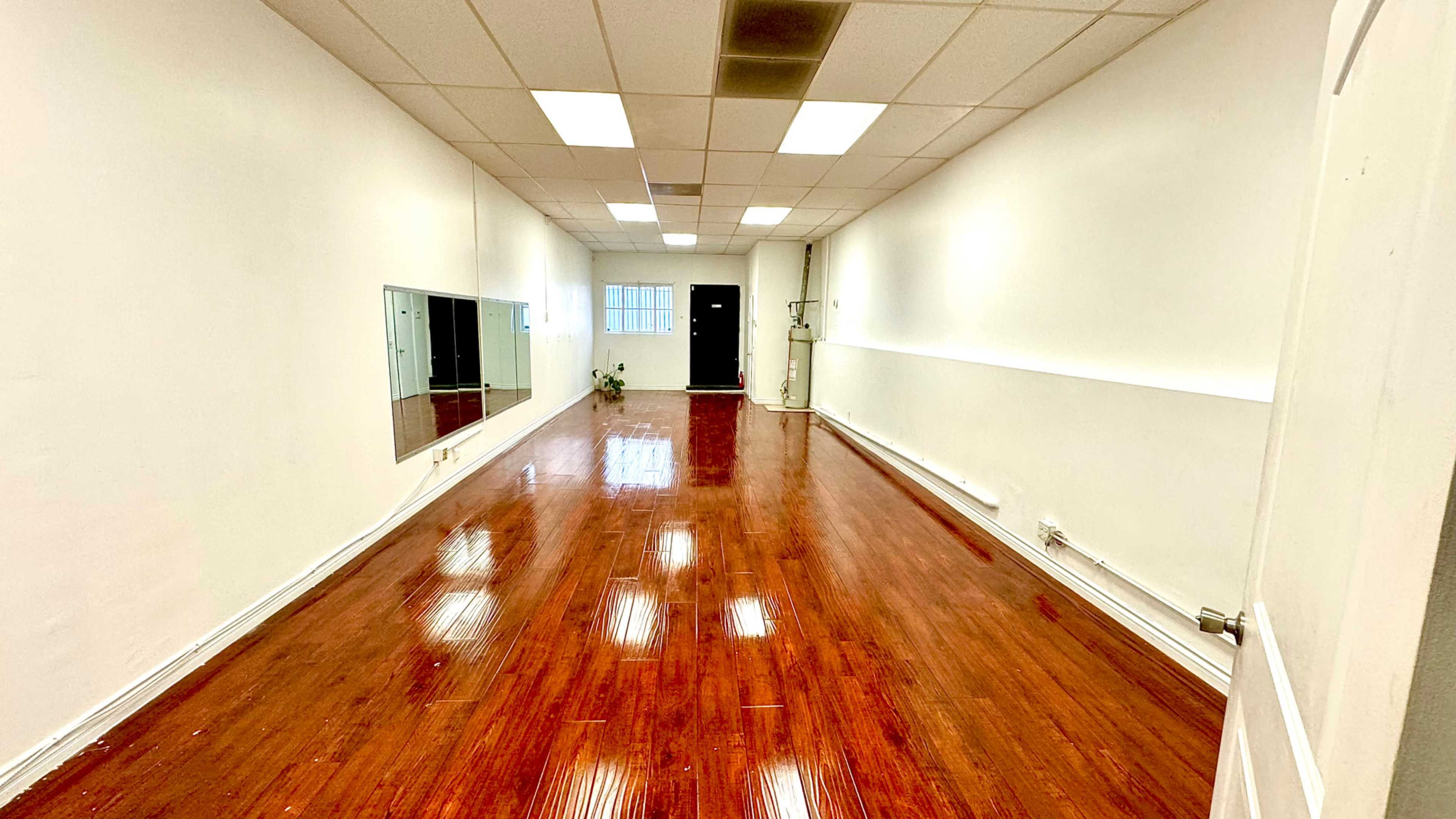 A spacious room with polished wooden floors, white walls, a large mirror on one side, and a door at the far end.